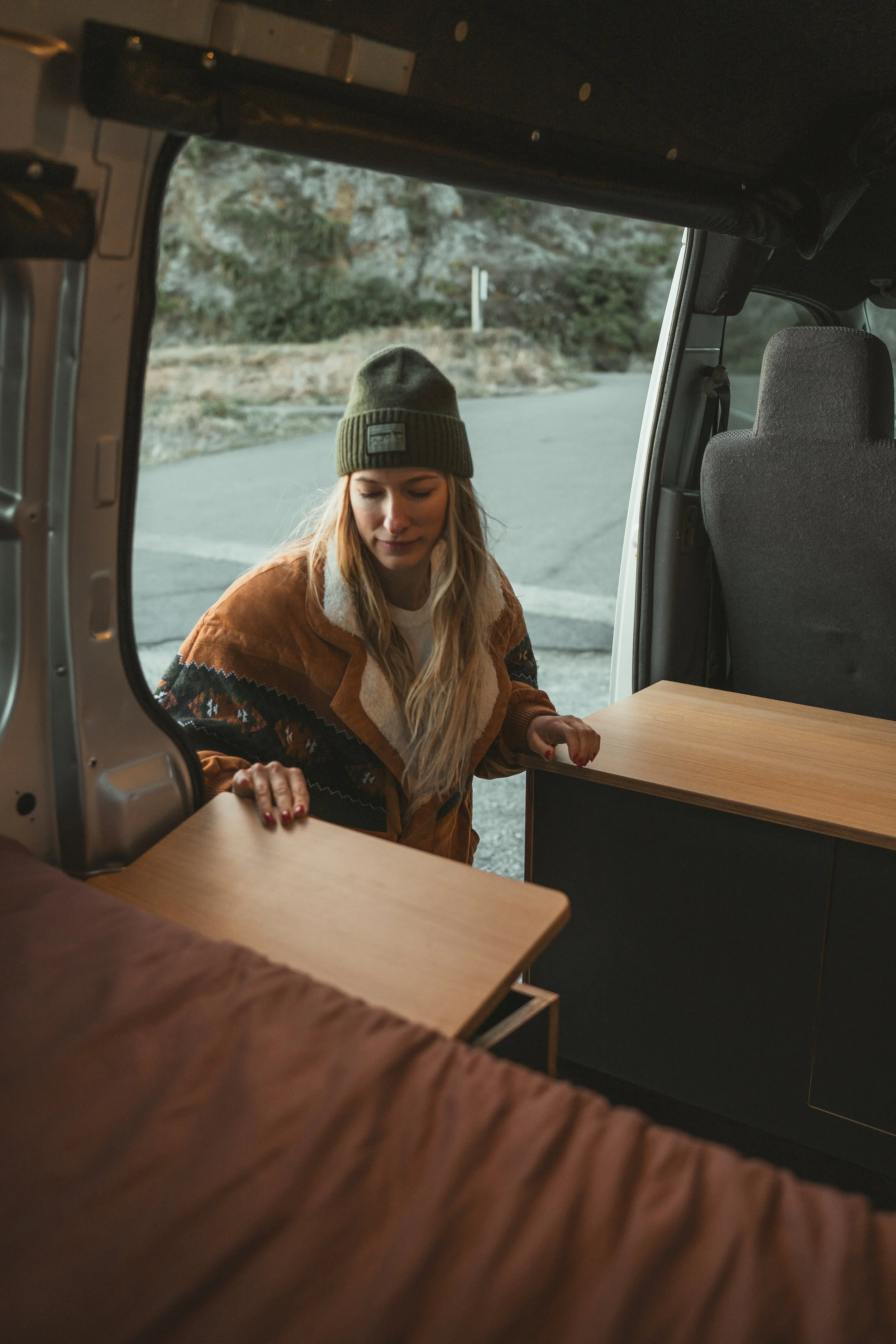 A woman with long blonde hair wearing a beanie and a patterned brown jacket loading a wooden table into the back of an open van parked outdoors.