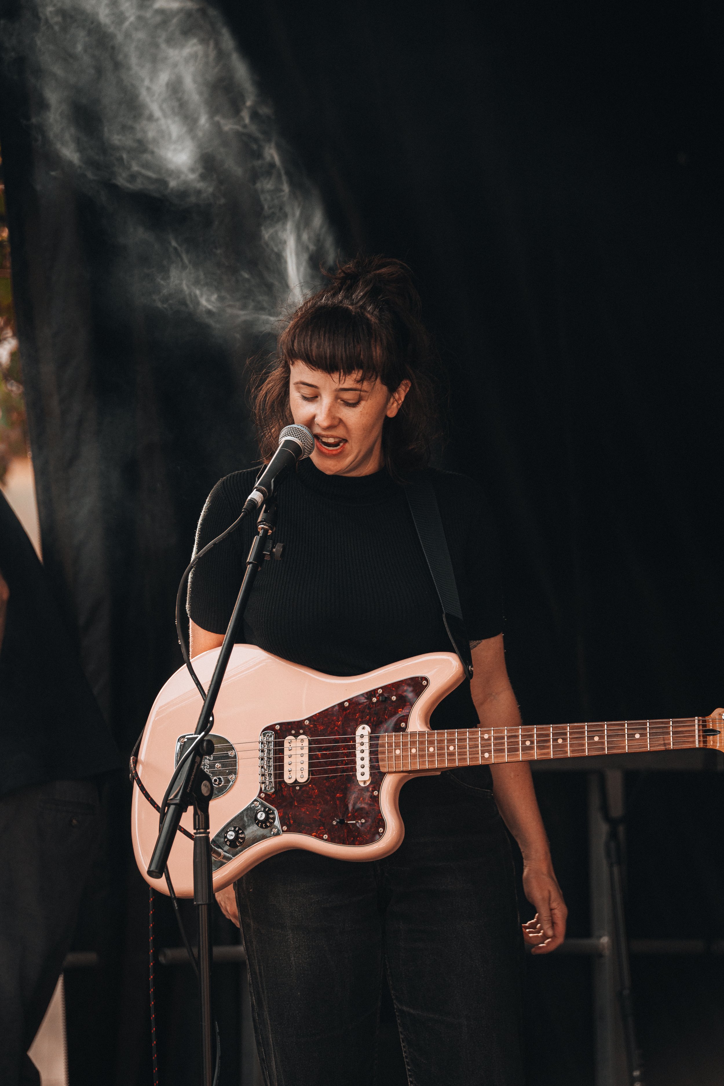 A woman with dark hair and bangs singing into a microphone while playing a pink electric guitar with a paisley pickguard, standing in front of a black background.