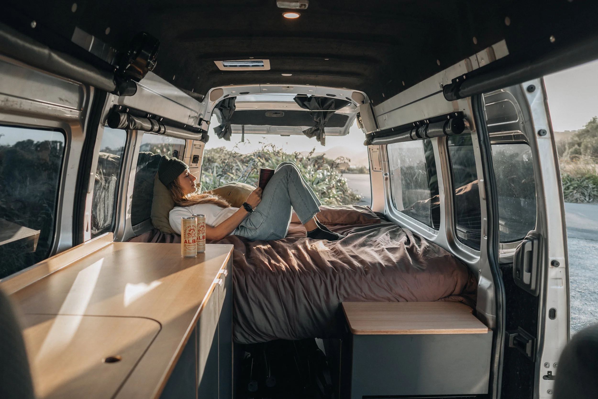 A person wearing a beanie and casual clothes is lying on a bed inside a camper van, reading a book. There's a can of ALB beer on a small table beside the bed, with natural light coming through the large windows, suggesting a relaxed outdoor setting.