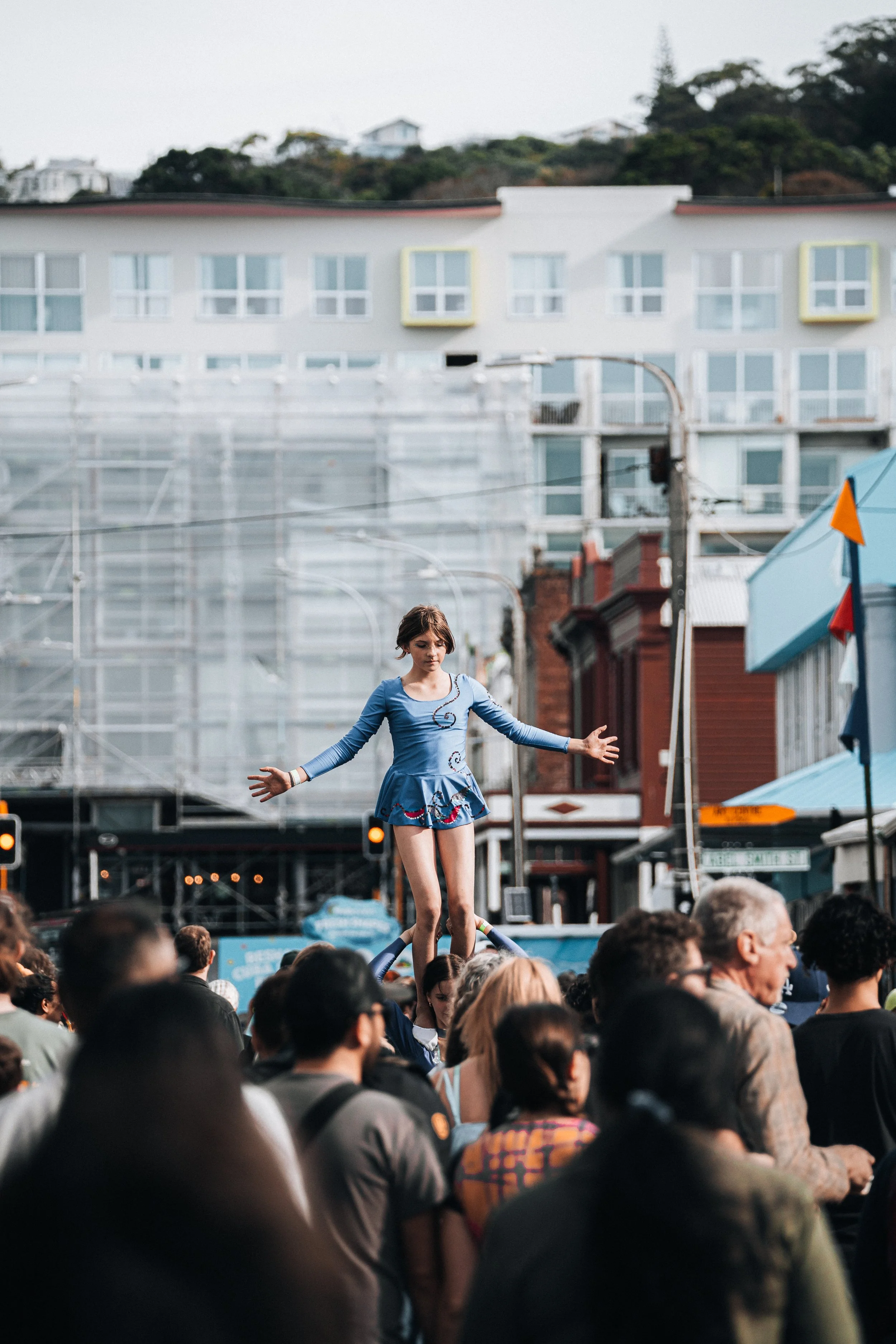 A young woman in a blue dress practicing acrobatics on a tightrope above a crowd during a street festival.