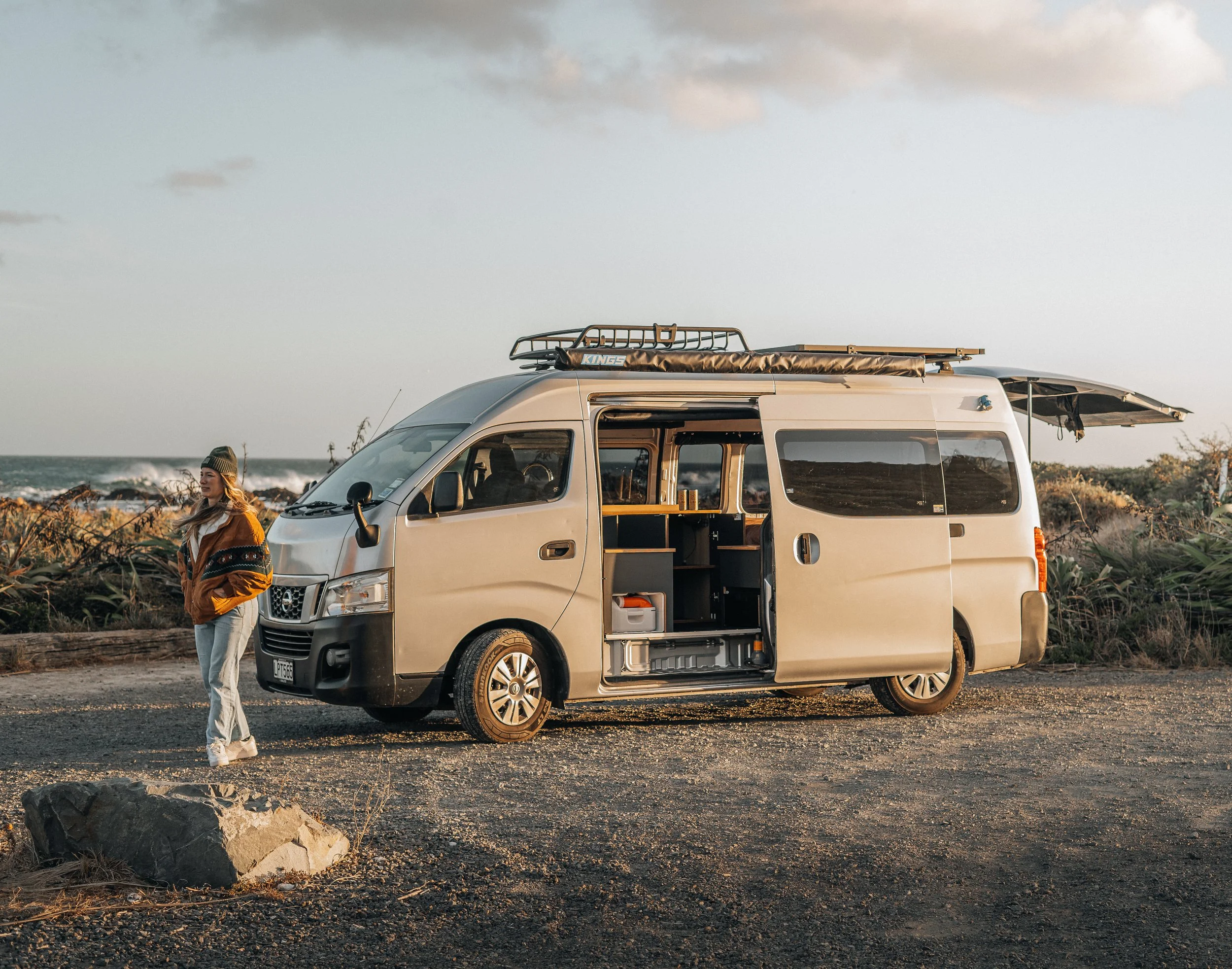 A woman standing outside a beige camper van parked on a dirt road near the ocean during sunset.