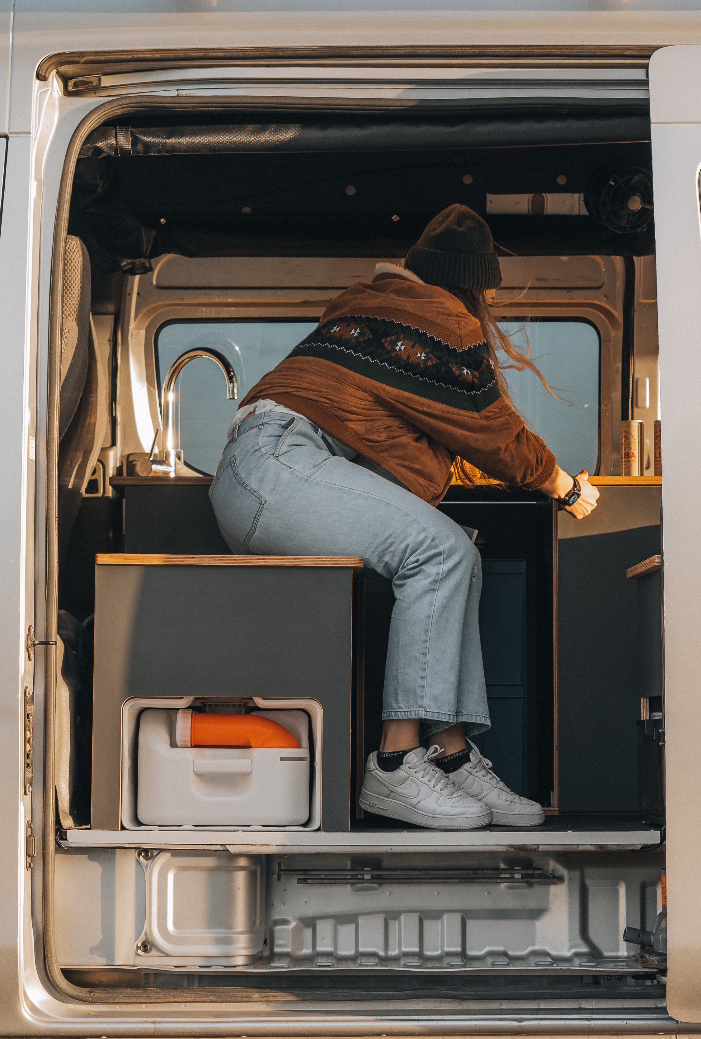 A person in a brown jacket, light blue jeans, white sneakers, and a dark beanie is inside a compact camper van, reaching for something on the countertop, with a small sink visible nearby.