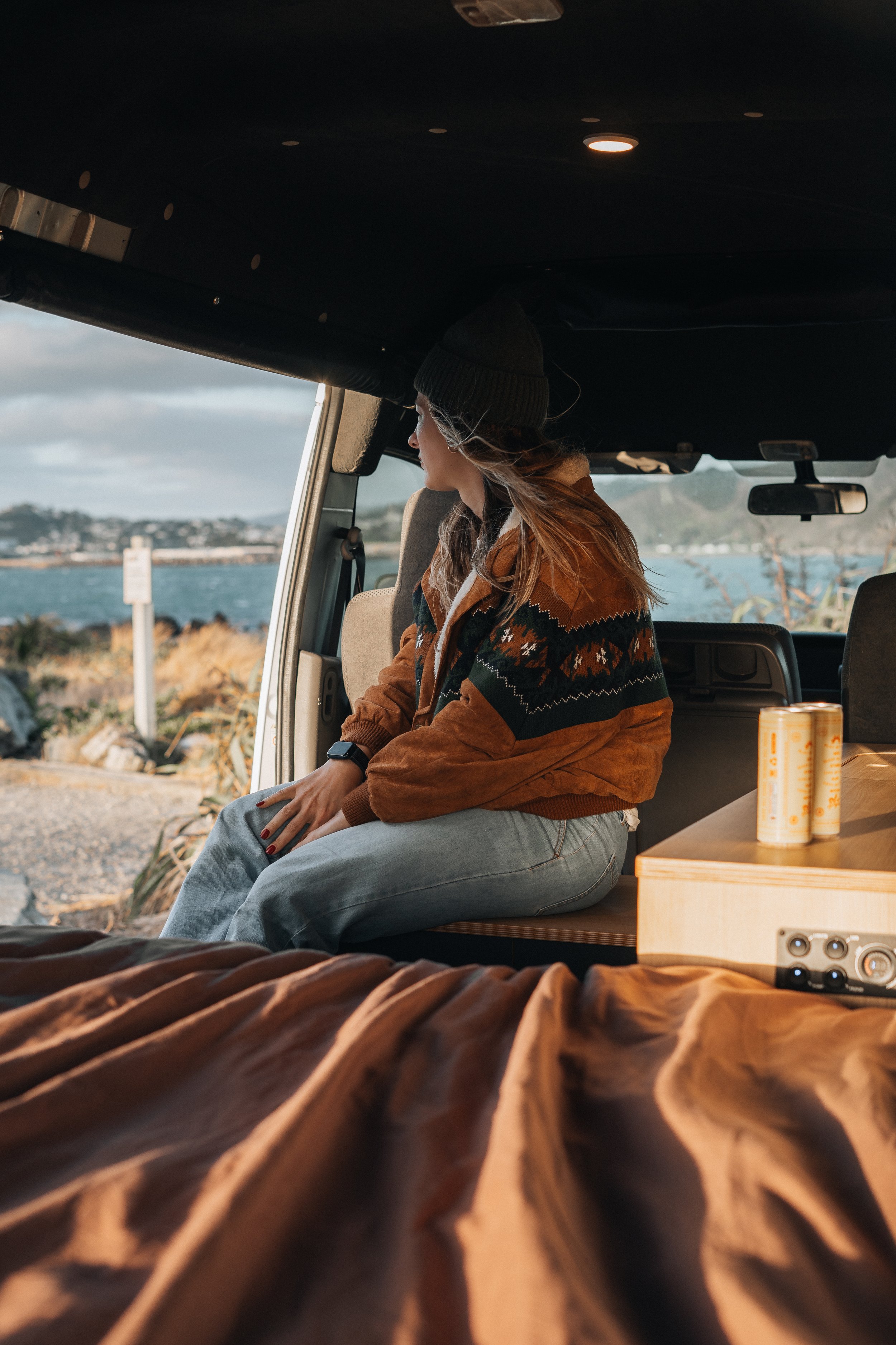 Woman sitting inside a camper van, looking out at a body of water with a coastline in the distance, wearing a beanie and patterned jacket.