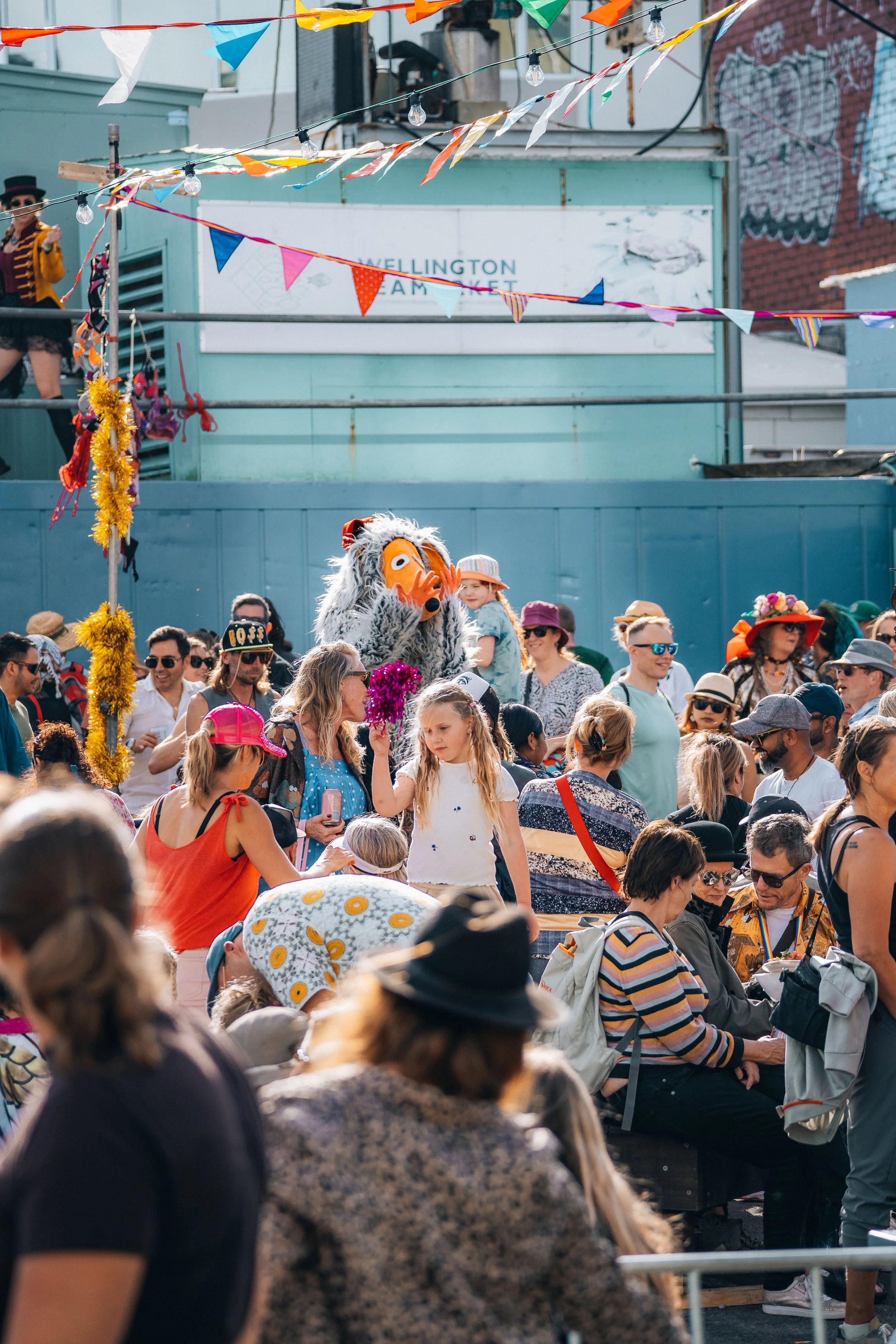 Crowd of people at an outdoor festival with colorful decorations and string lights, including a person dressed as a lion mascot.