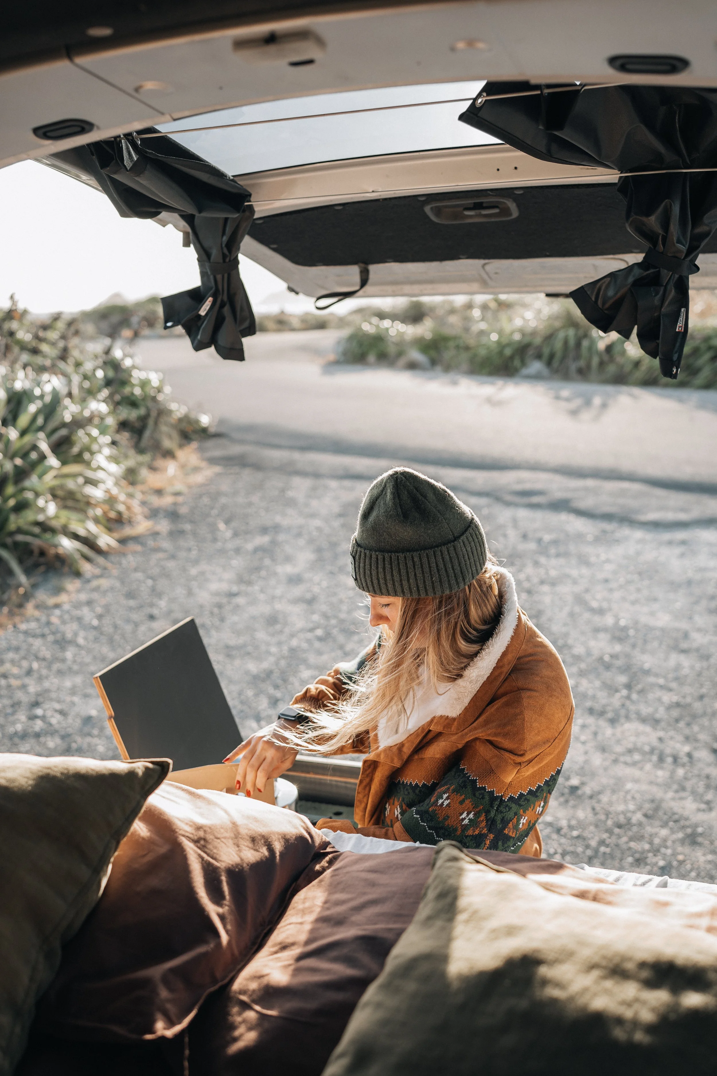 A woman sitting on the ground behind an open trunk of a car, looking at a book, on a scenic outdoor road.