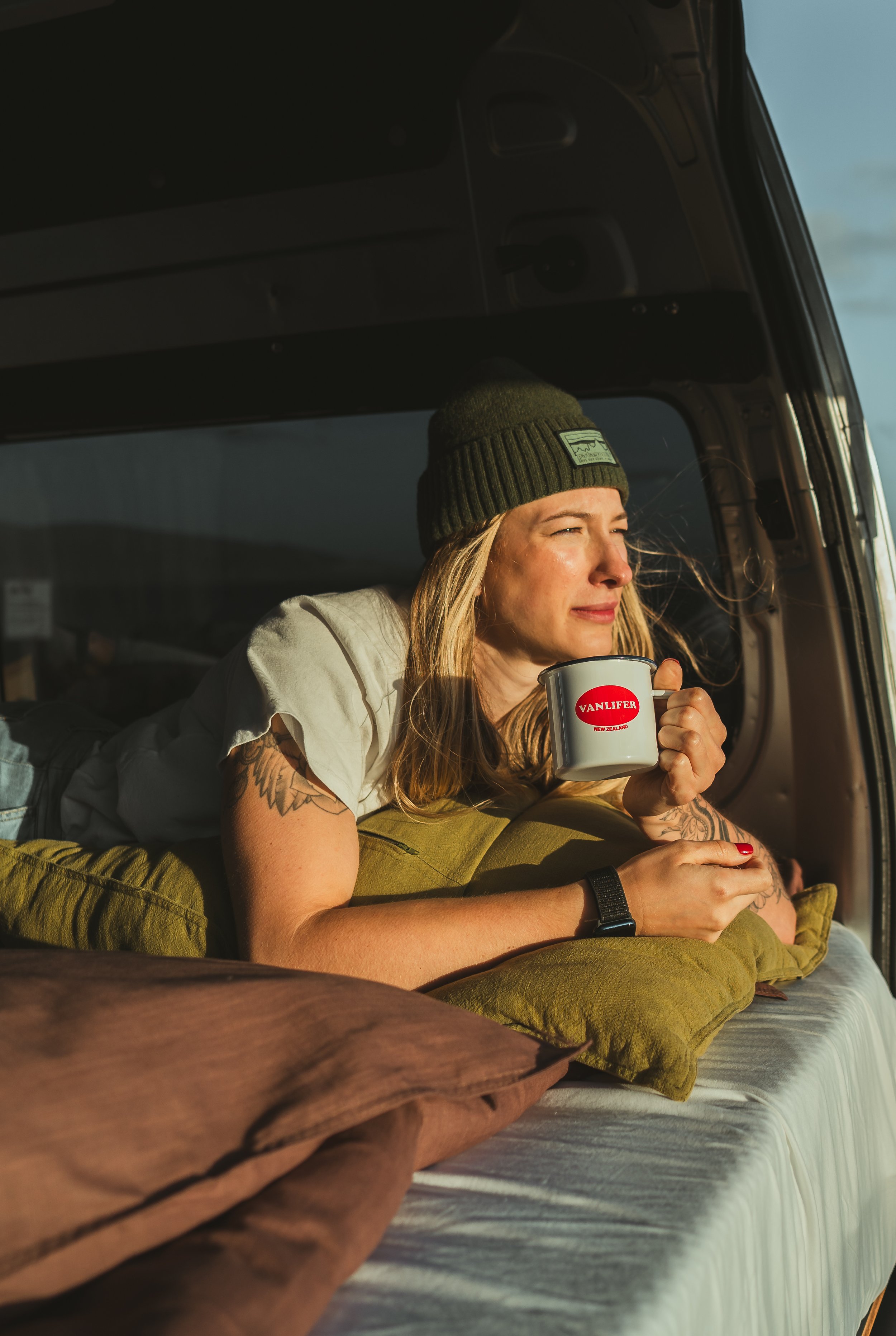 A woman with tattoos and wearing a beanie hat relaxes inside a van, lying on cushions and holding a mug with the Vanliner logo, gazing out the window at sunset.