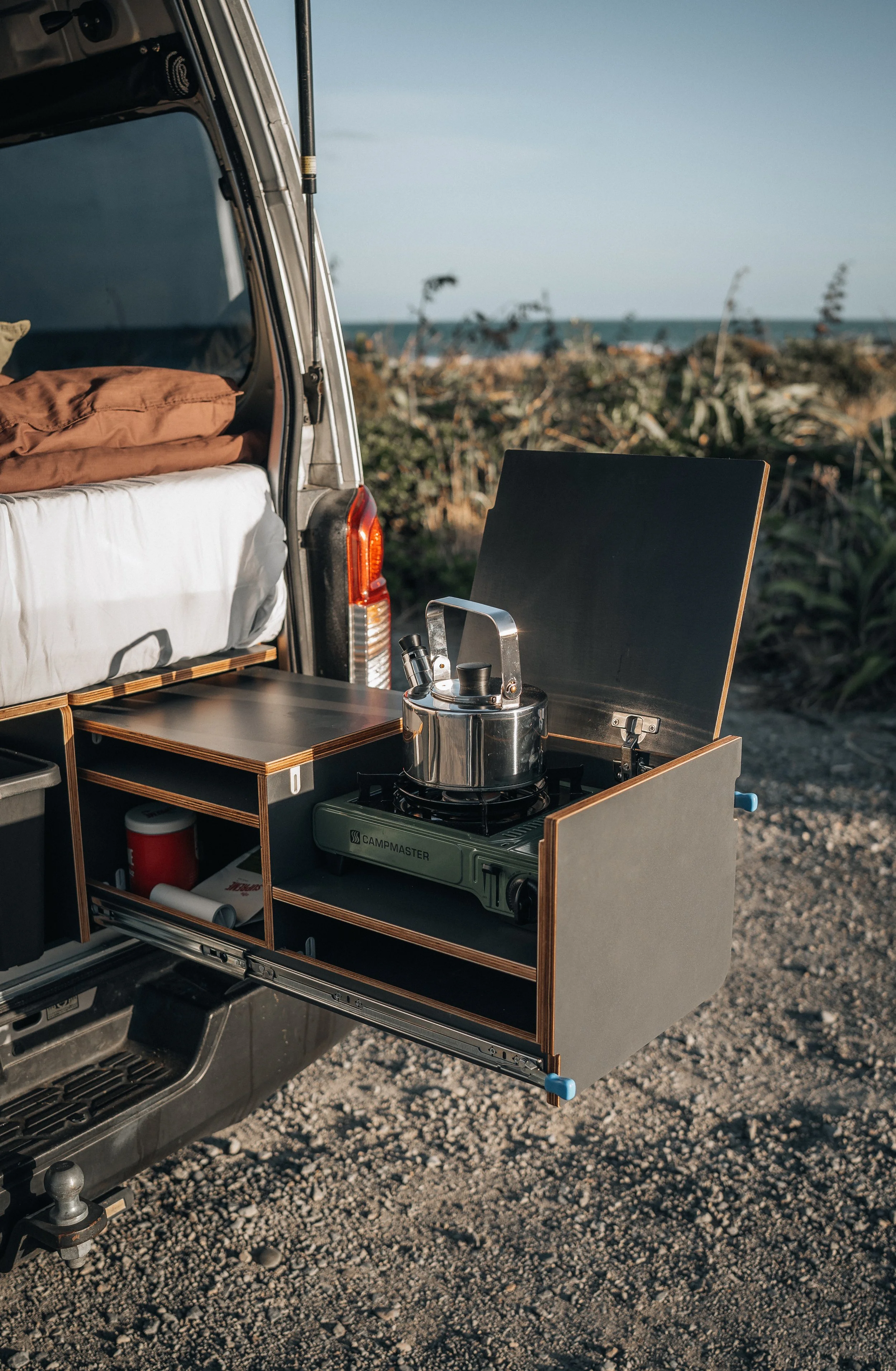 Open camper van showing a portable stove with a kettle, set up on a fold-out storage drawer next to a bed, with a coastal landscape in the background.