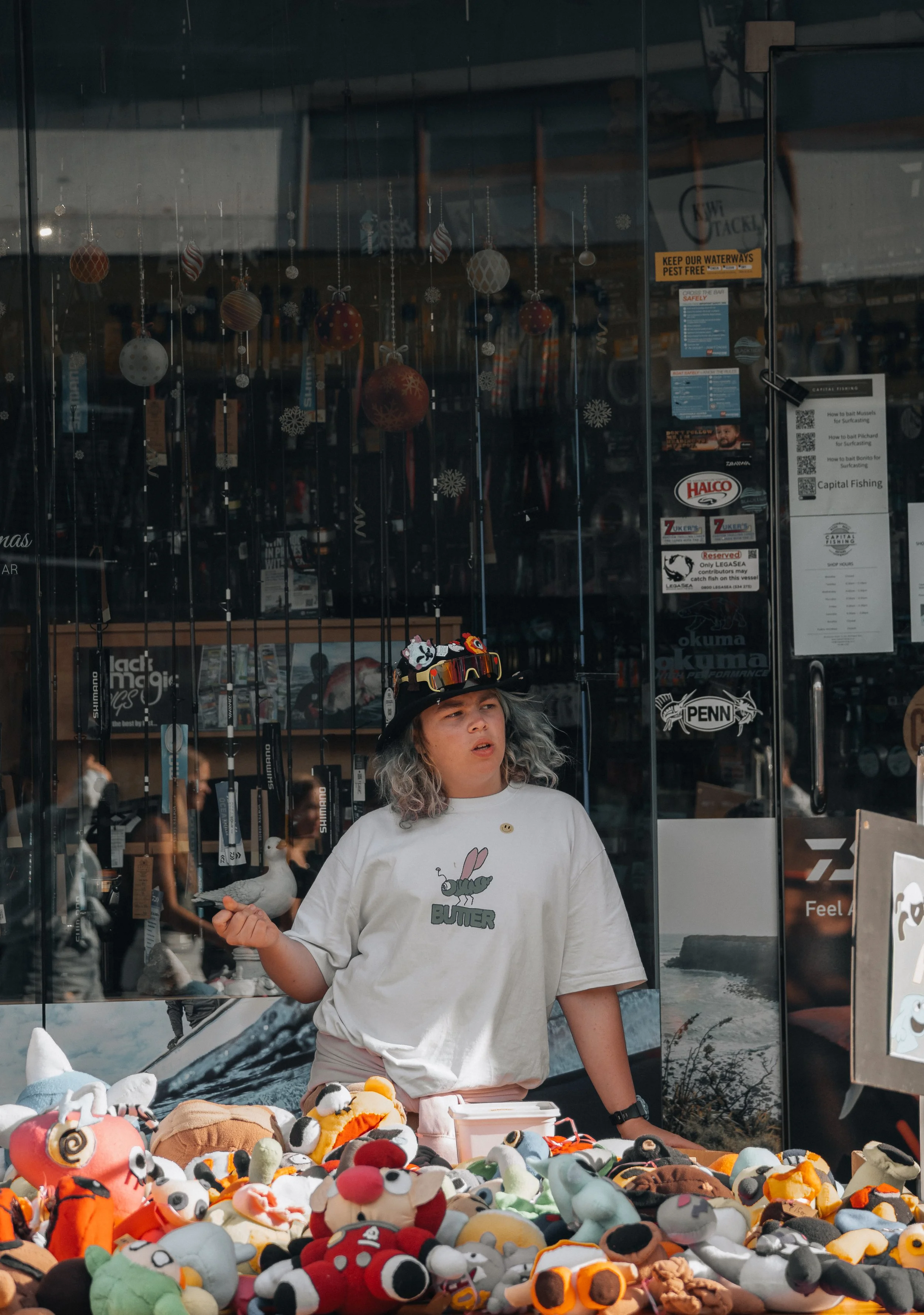 A woman with gray curly hair wearing a white t-shirt with a bunny and snail graphic and the word 'BUTTER' on it, standing behind a table filled with colorful plush toys and stuffed animals outside a store with fishing equipment visible through the gl
