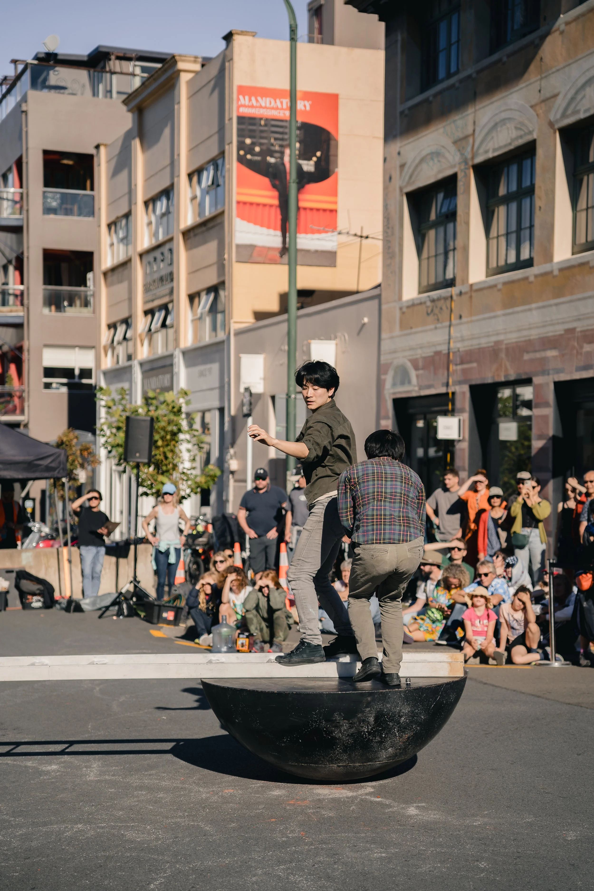Two performers on a black circular platform performing in front of a crowd on a city street. One person stands and appears to be in motion, while the other is kneeling or bending down. The audience is seated and standing, watching the performance in 
