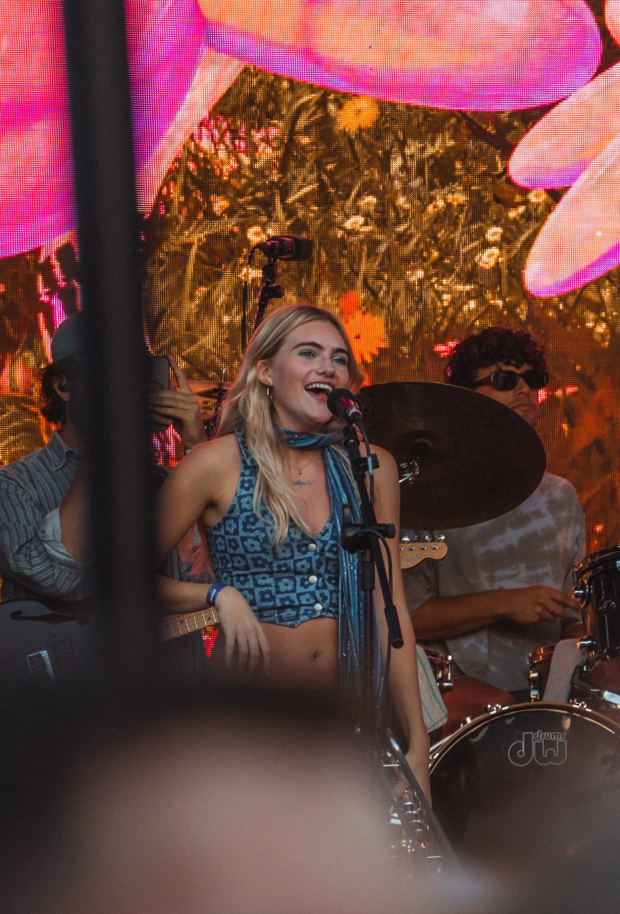 A young woman with long blonde hair singing into a microphone during a live performance, accompanied by band members playing drums and guitar, with a colorful digital backdrop behind them.