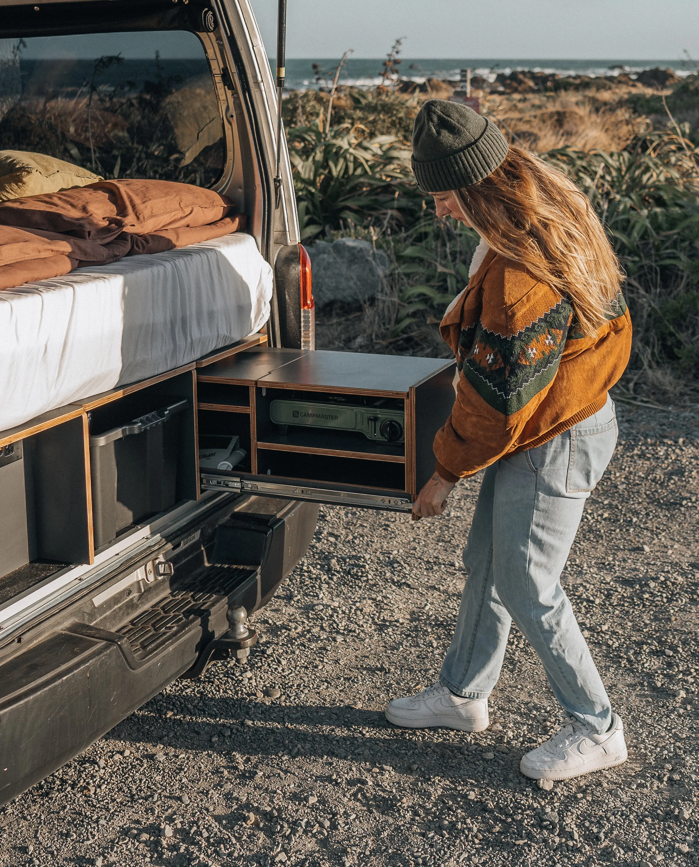 A woman in a brown jacket, gray beanie, and sneakers opening a storage drawer on the back of a camper van parked near the beach, with vegetation and the ocean in the background.
