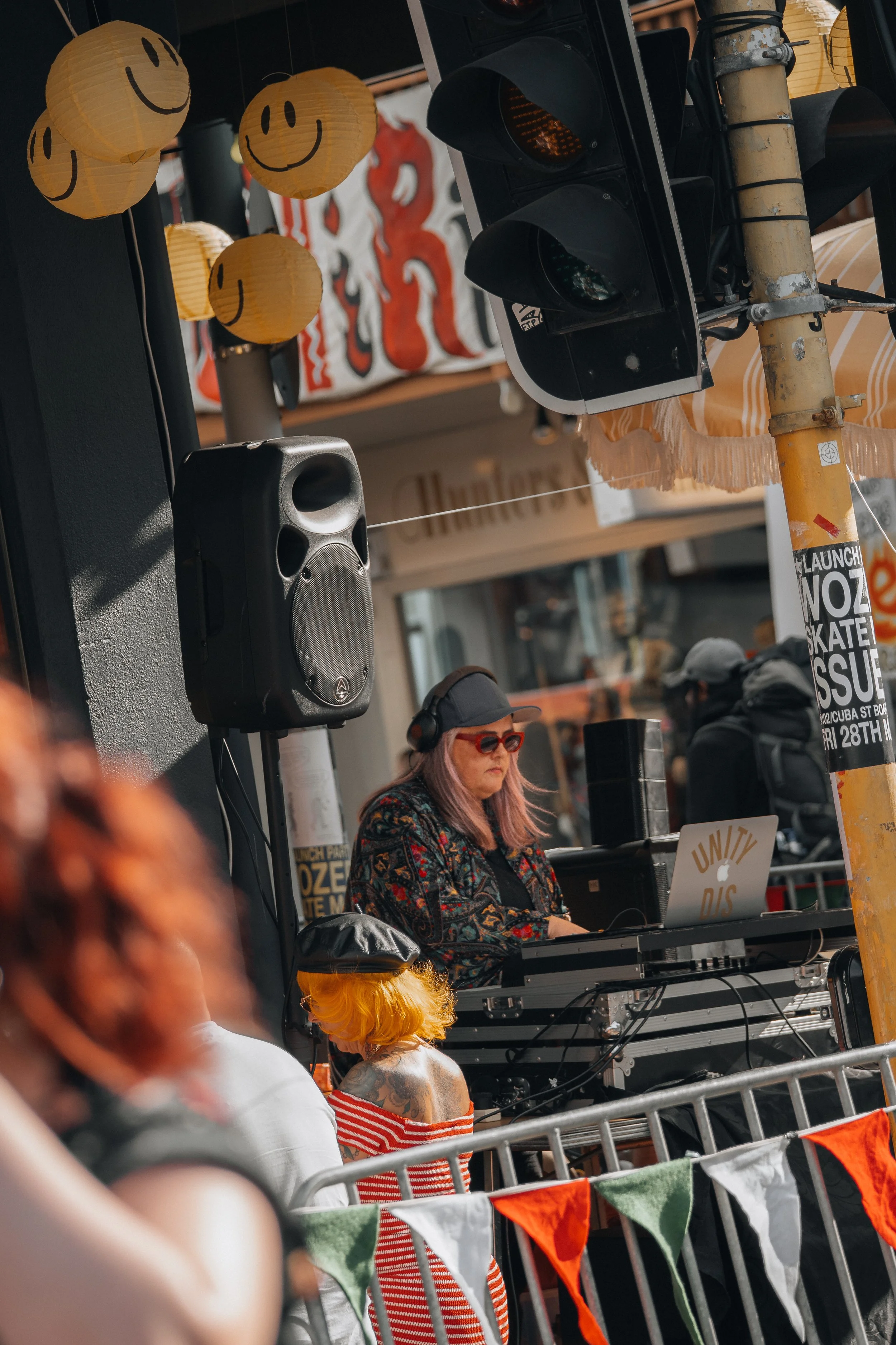 A DJ wearing sunglasses and headphones performs at an outdoor event with colorful decorations, including yellow paper lanterns with smiley faces, and a crowd of people in front.