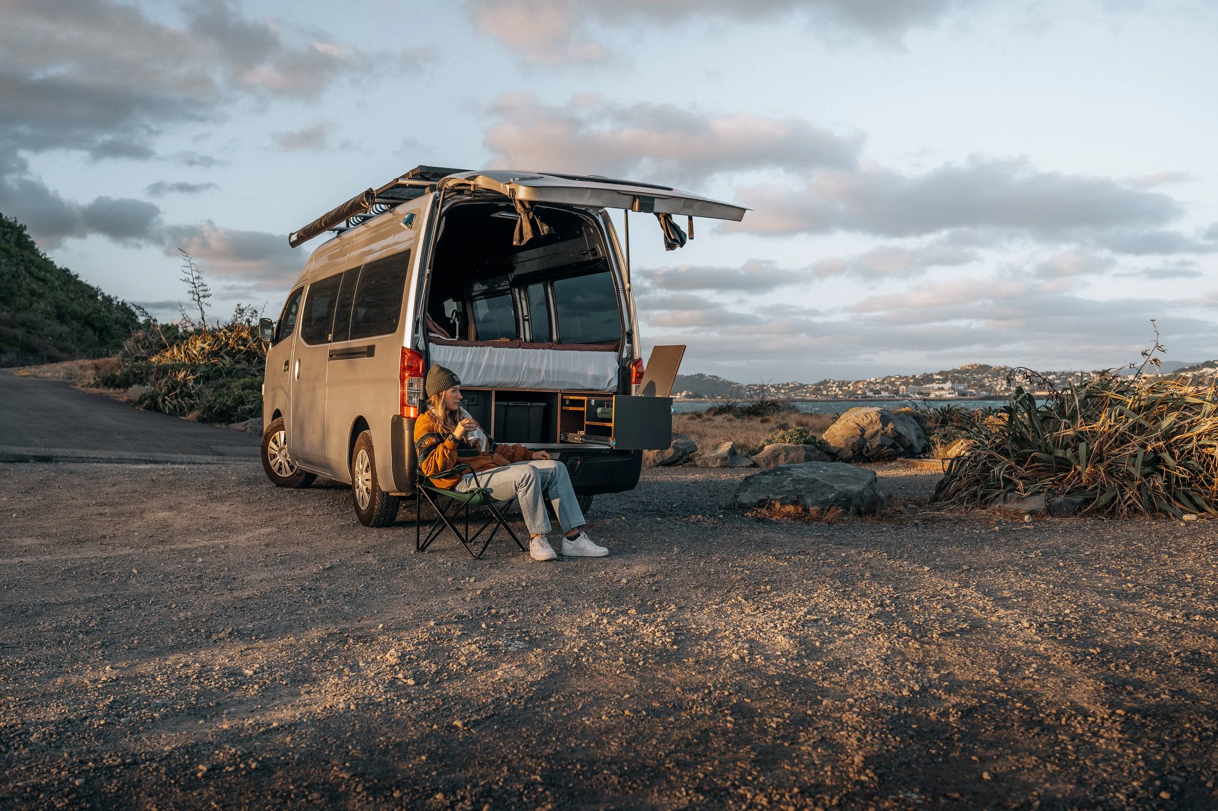 A woman sitting on a camping chair outside a van parked on a dirt road near a rocky coastline with hills and buildings in the distance, drinking from a cup during sunset or sunrise.