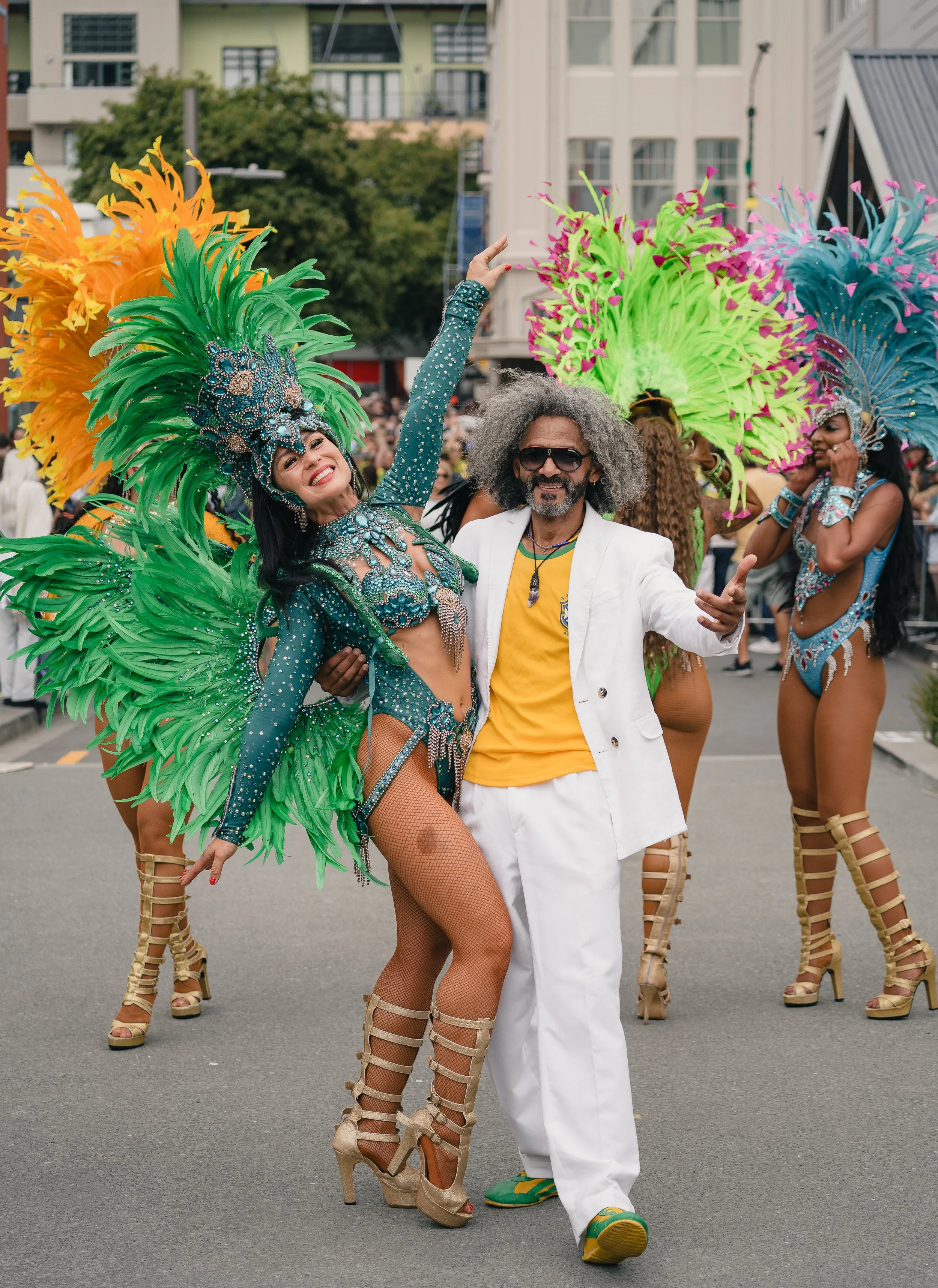 People in colorful carnival costumes dancing outdoors during a parade, with a woman in a blue and green feathered headdress and costume, posing with a man in a white suit and sunglasses.