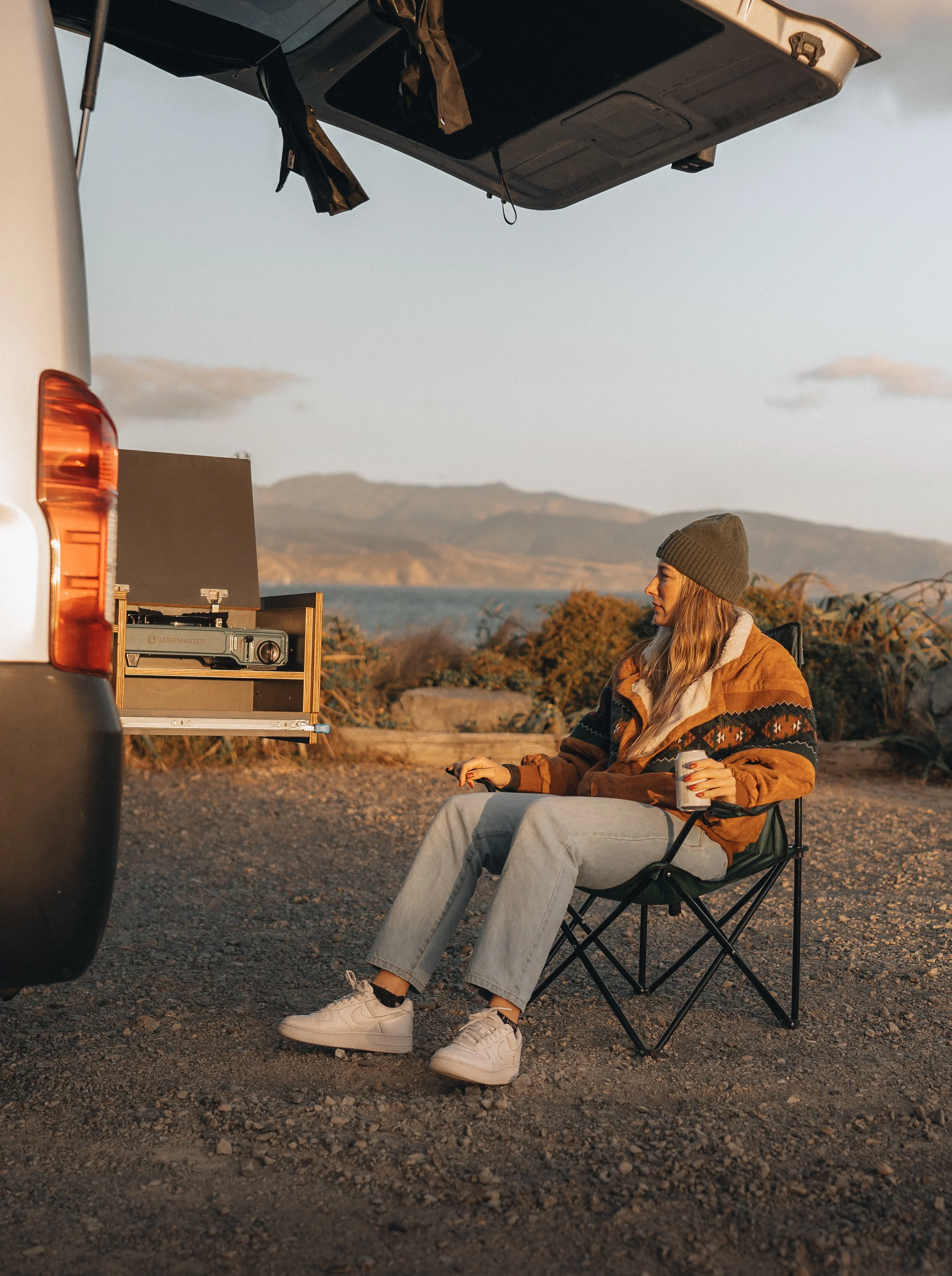 A woman sitting in a camping chair outside a vehicle, holding a drink, with a view of mountains and water in the background during sunset.