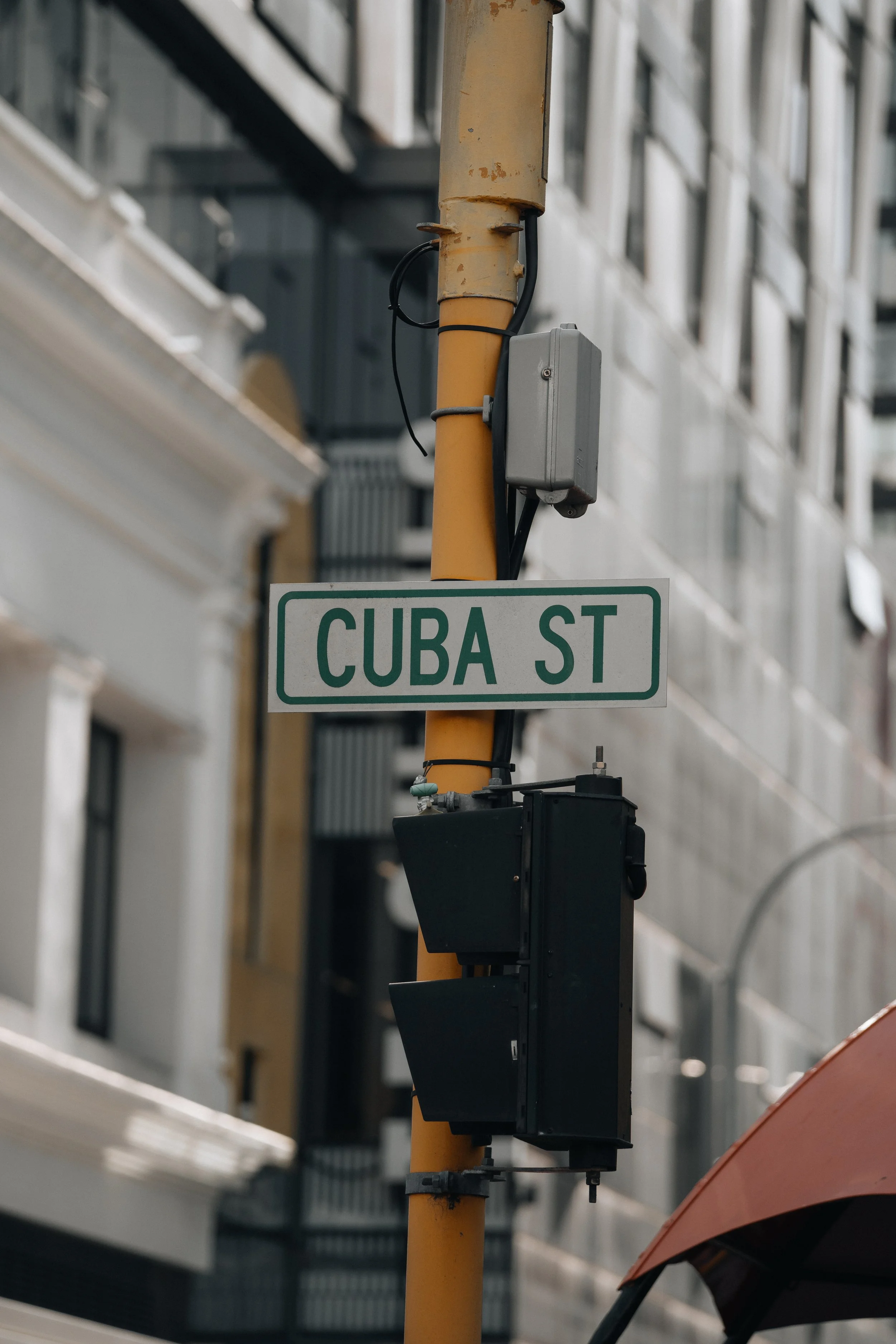 Street sign reading 'Cuba St' attached to a yellow pole with electrical components, against a background of urban buildings.