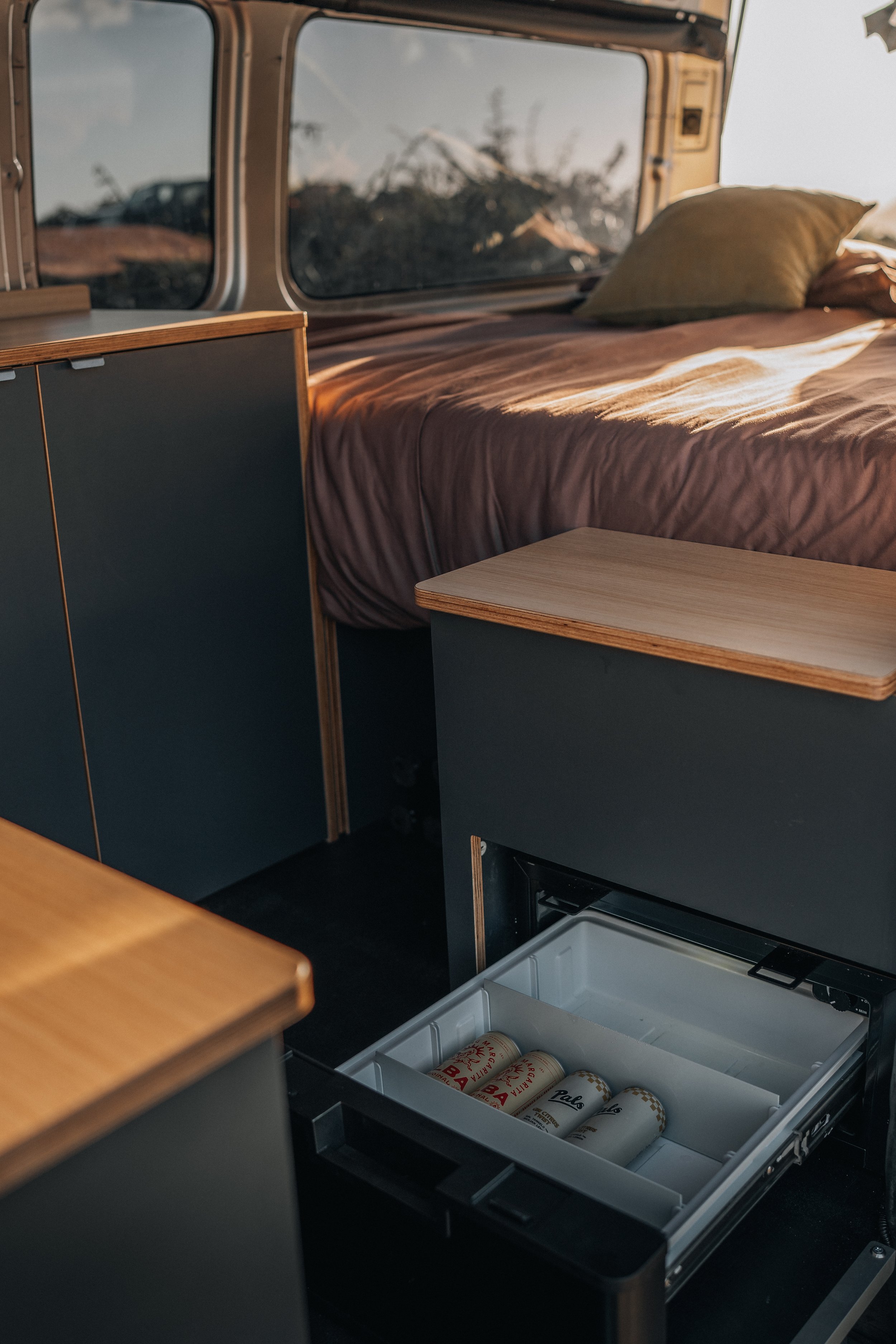 Interior of a camper van with a bed, small cabinets, and an open mini fridge containing cans of beer and soda.
