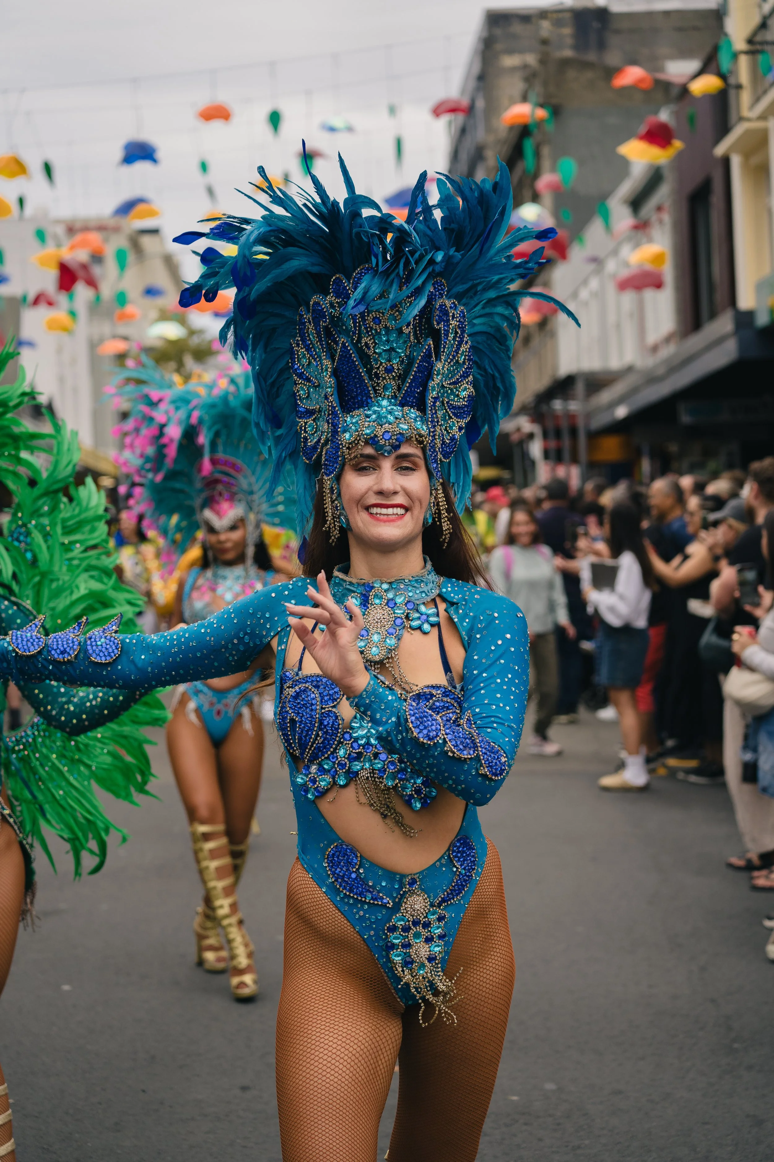 A woman in a vibrant blue carnival costume with feathers and rhinestones, smiling during a parade on a city street, with other dancers and spectators in the background.