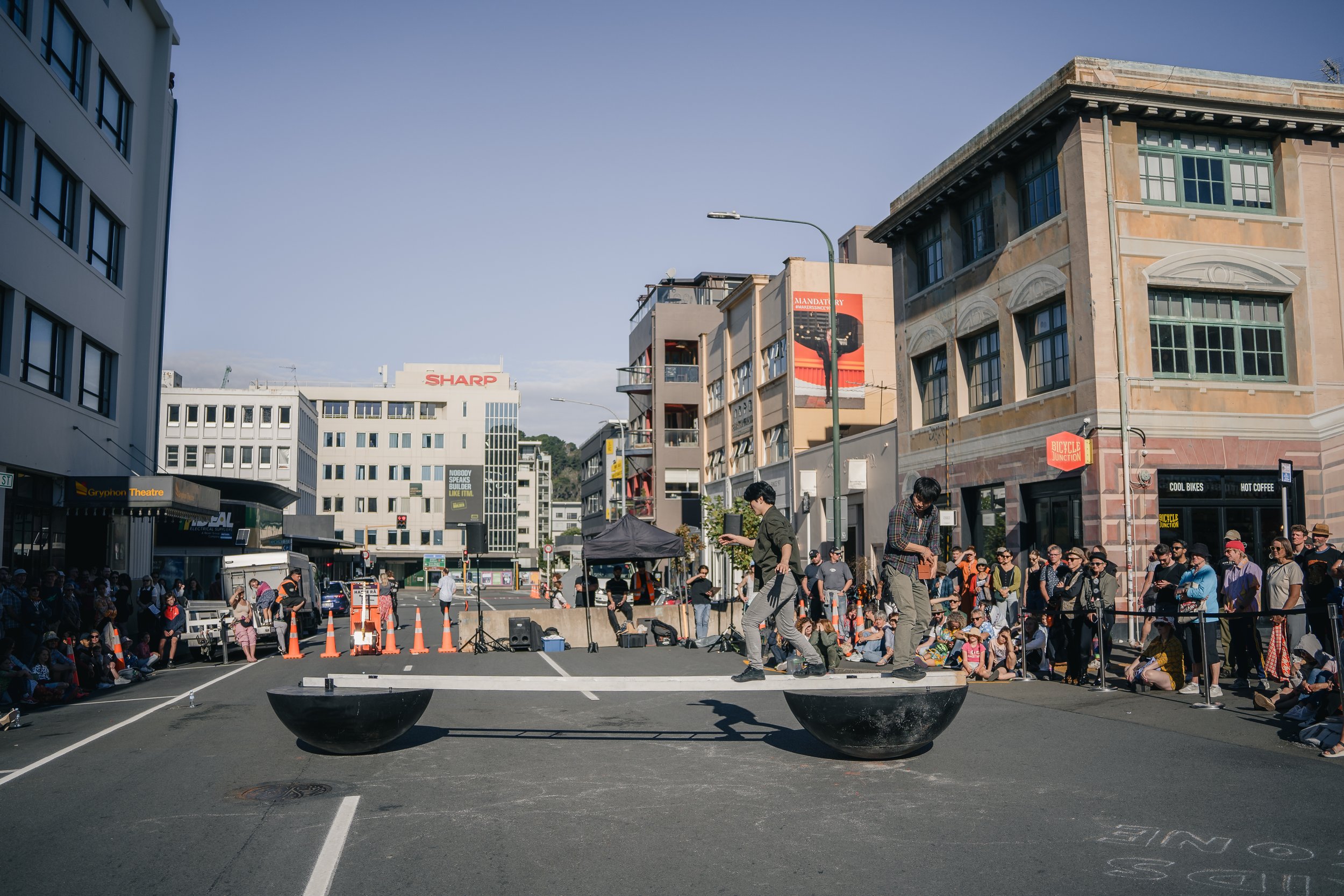 Two performers walking on suspended bowls in the middle of a street surrounded by a seated crowd and onlookers, with city buildings in the background.