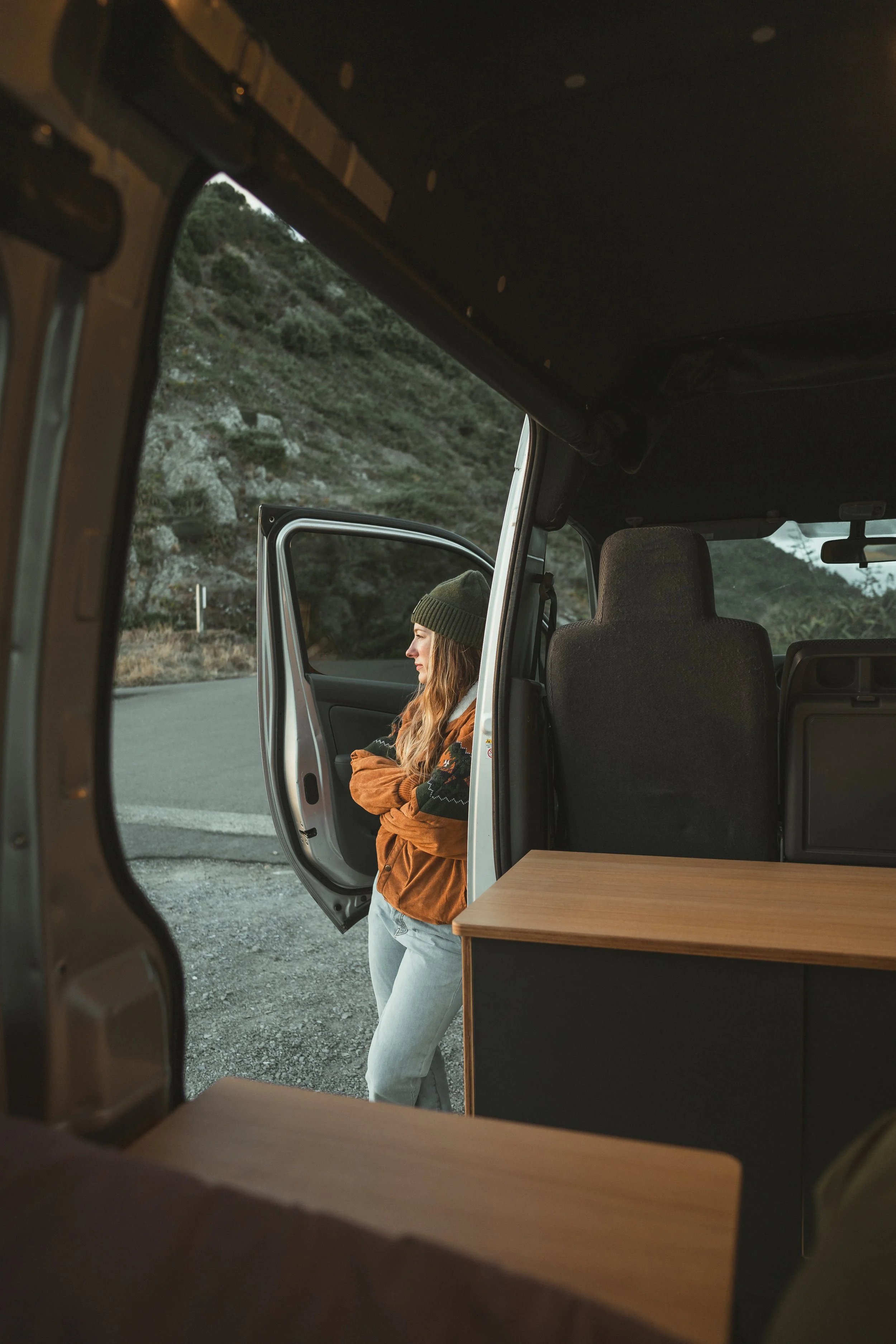 A woman standing outside a van with her arms crossed, looking to the side, wearing a green beanie, a brown jacket, and light blue jeans, in a mountainous area near a road.