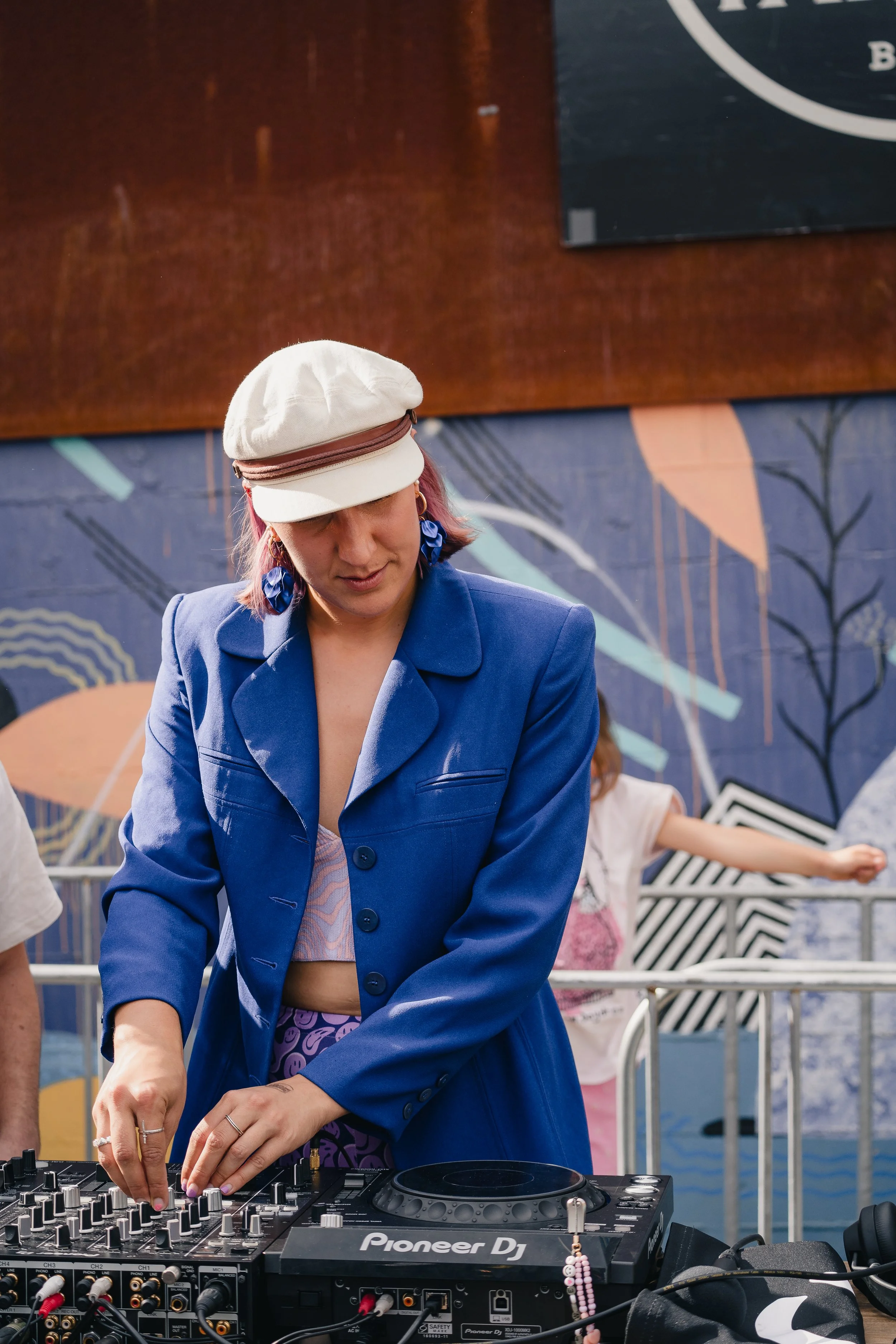 A woman DJ wearing a white cap and a blue blazer, mixing music at a DJ console in front of a colorful abstract mural.