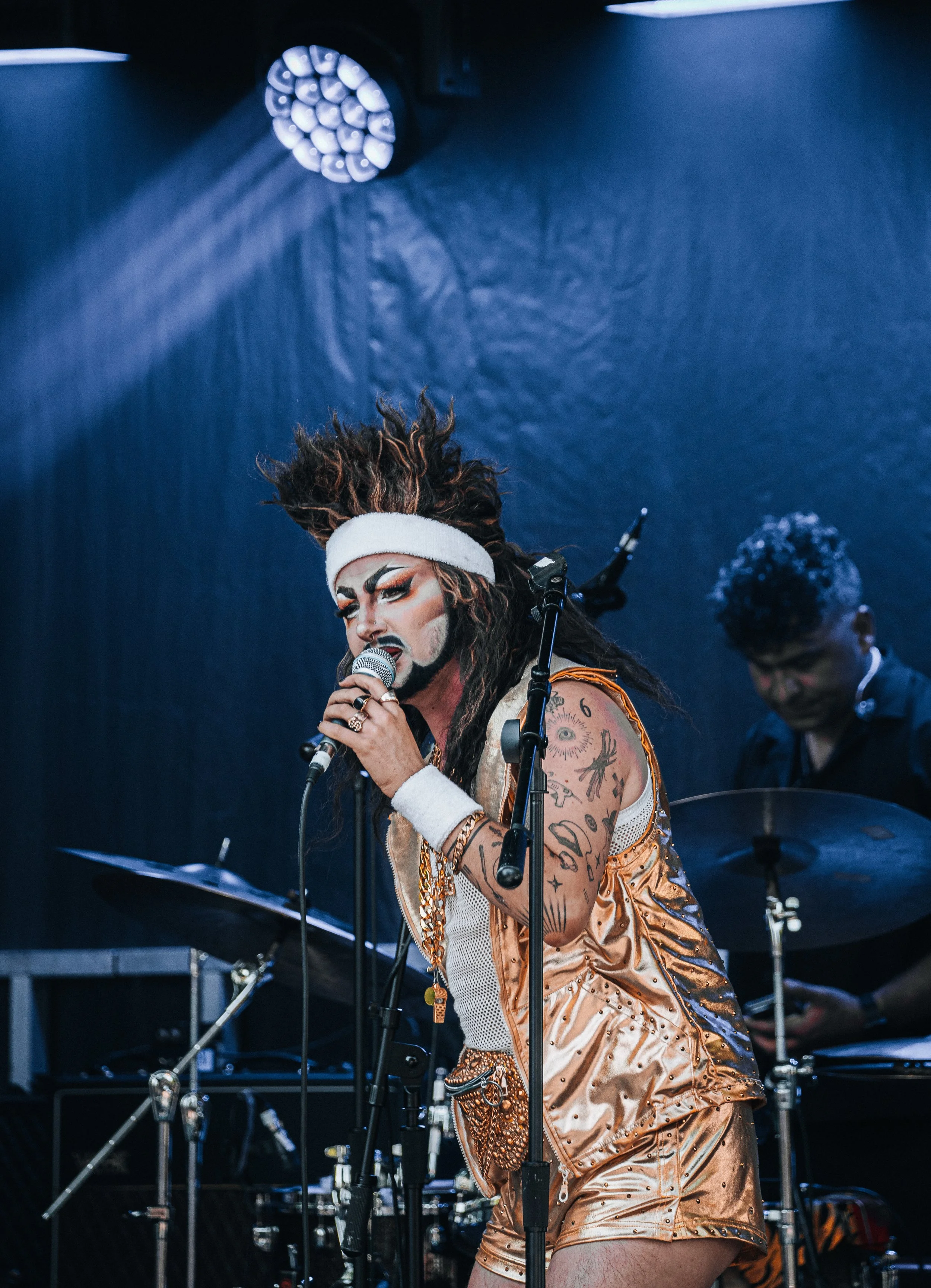 A performer with dreadlocks, painted face, and Eastern-inspired costume singing into a microphone on stage, with a drummer playing in the background.