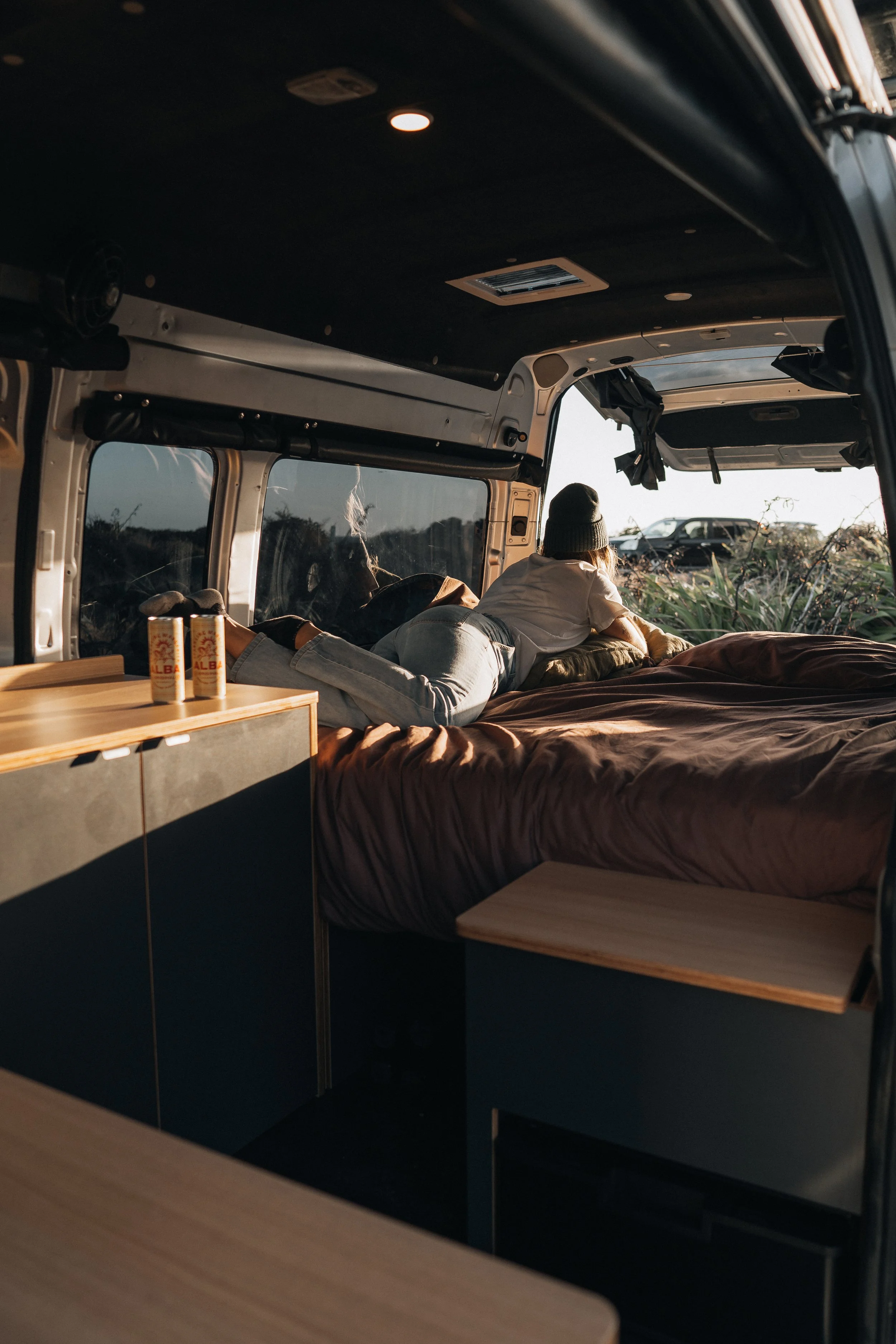 A person lying on a bed inside a camper van, looking out the window at a sunset or sunrise in a natural setting with a car parked outside.