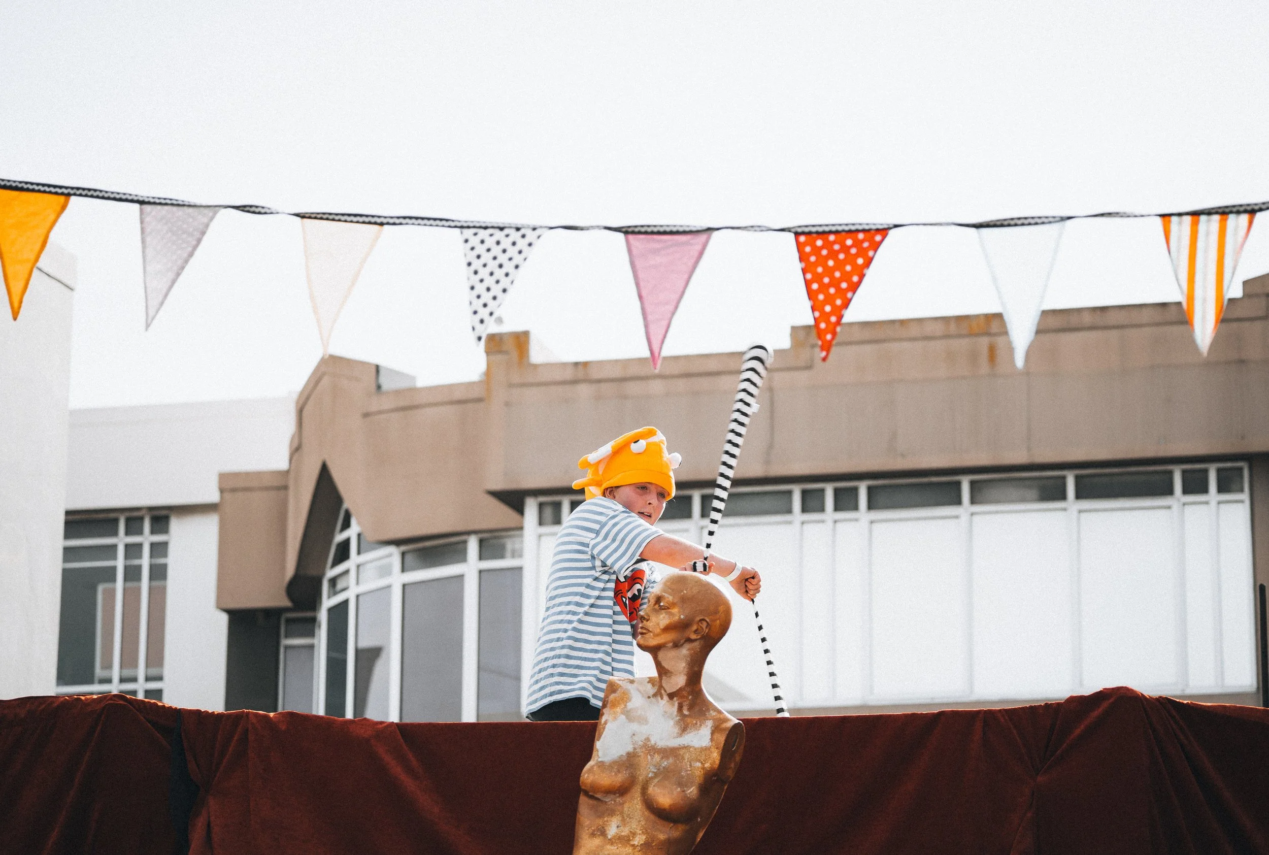 Child wearing a yellow hat and striped shirt, holding a striped stick, standing behind a statue of a woman, with colorful bunting flags hanging overhead, in front of a building.
