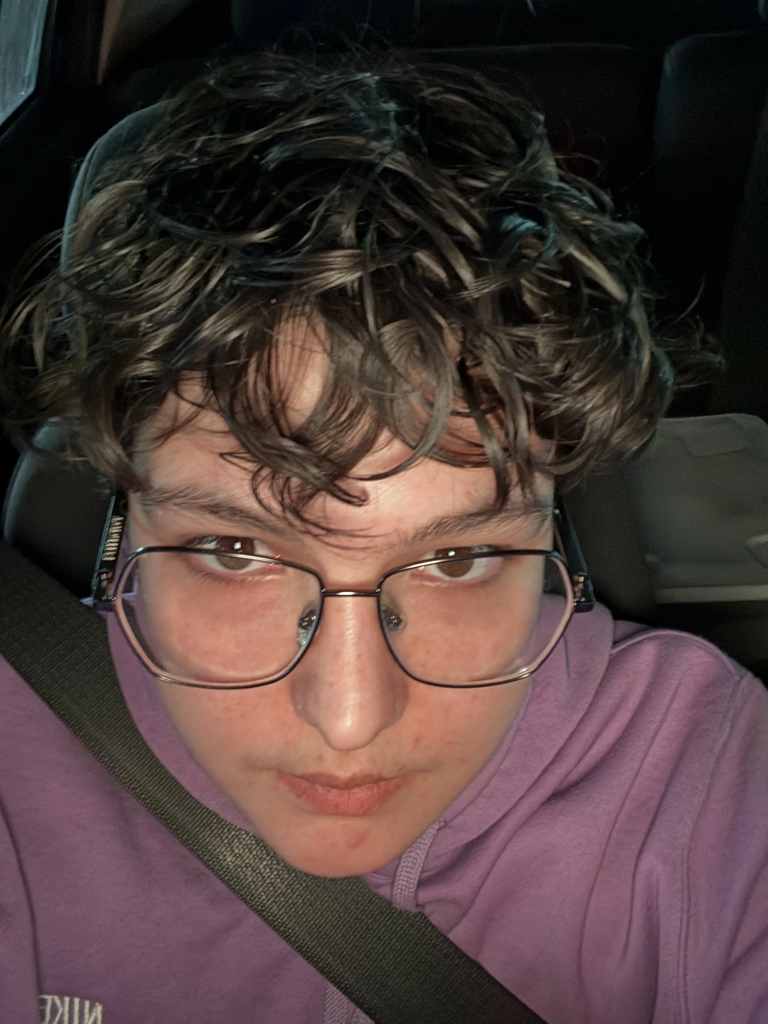 Close-up selfie of a person with curly hair wearing glasses and a purple hoodie, sitting in a vehicle.
