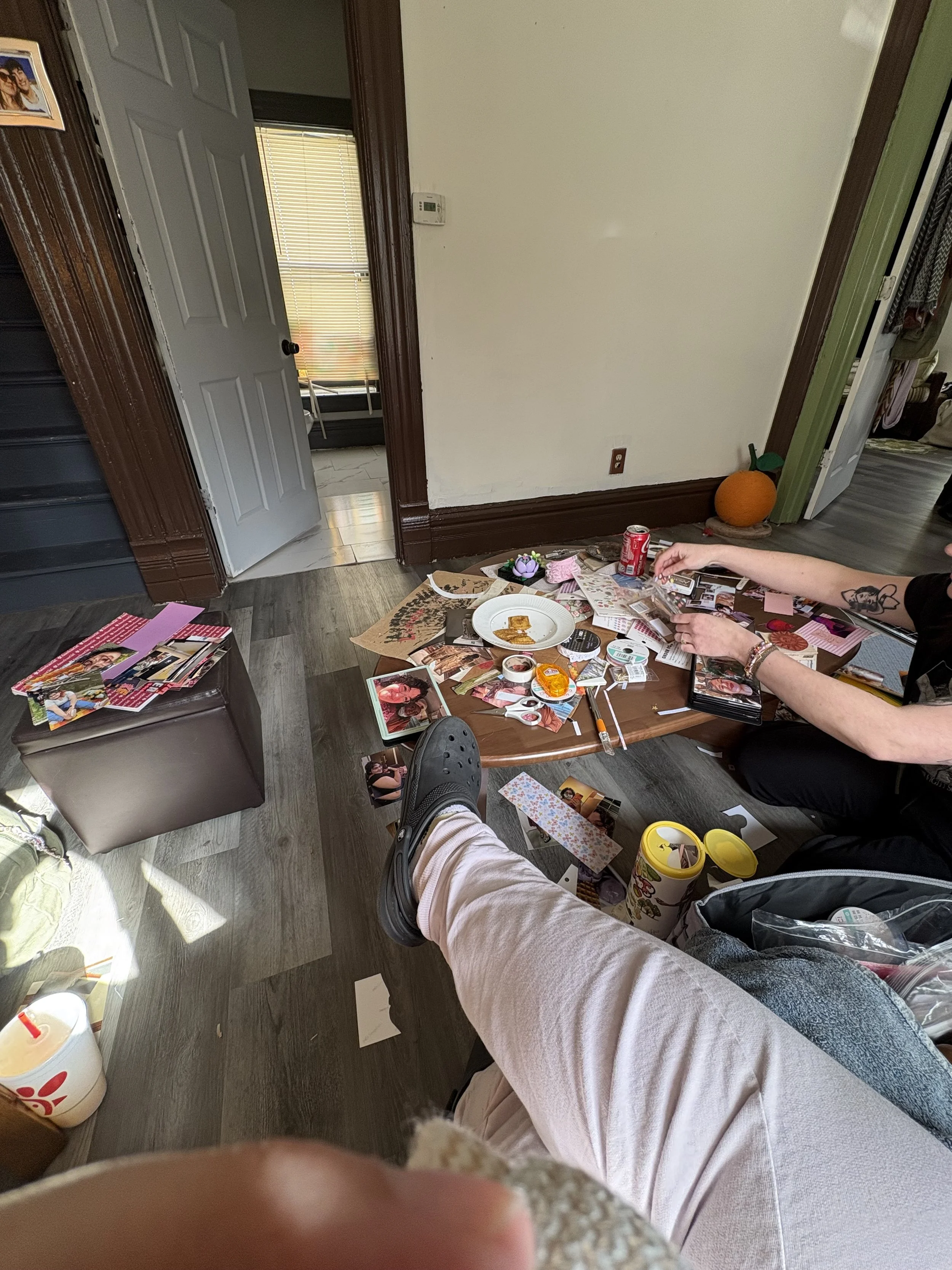 Messy coffee table with photos, snacks, and drinks in a living room, with a person’s legs and feet visible in the foreground, and a doorway and pumpkin decoration in the background.