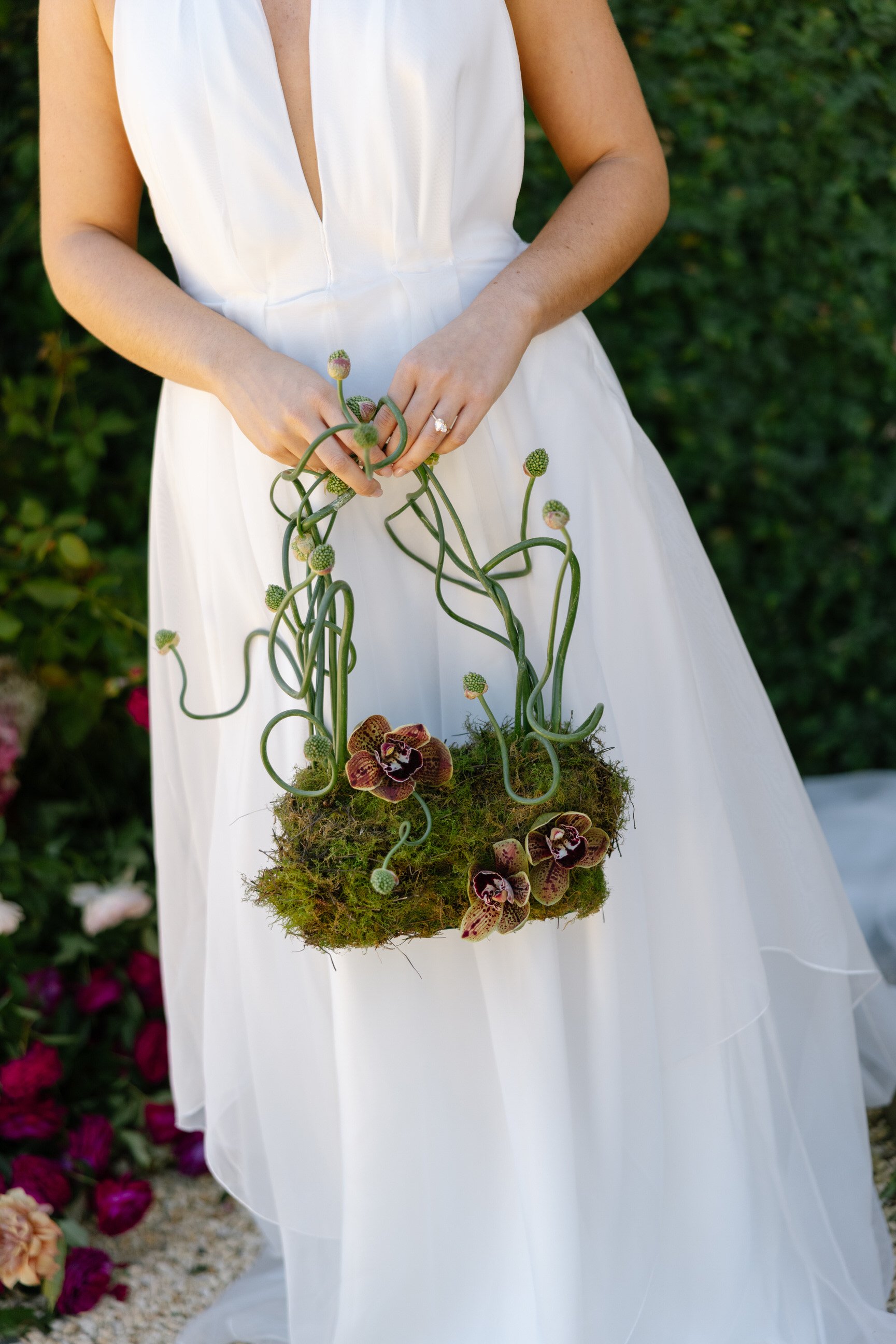 A woman in a white dress holding a floral arrangement with green vines, moss, and purple orchids against a background of green foliage.