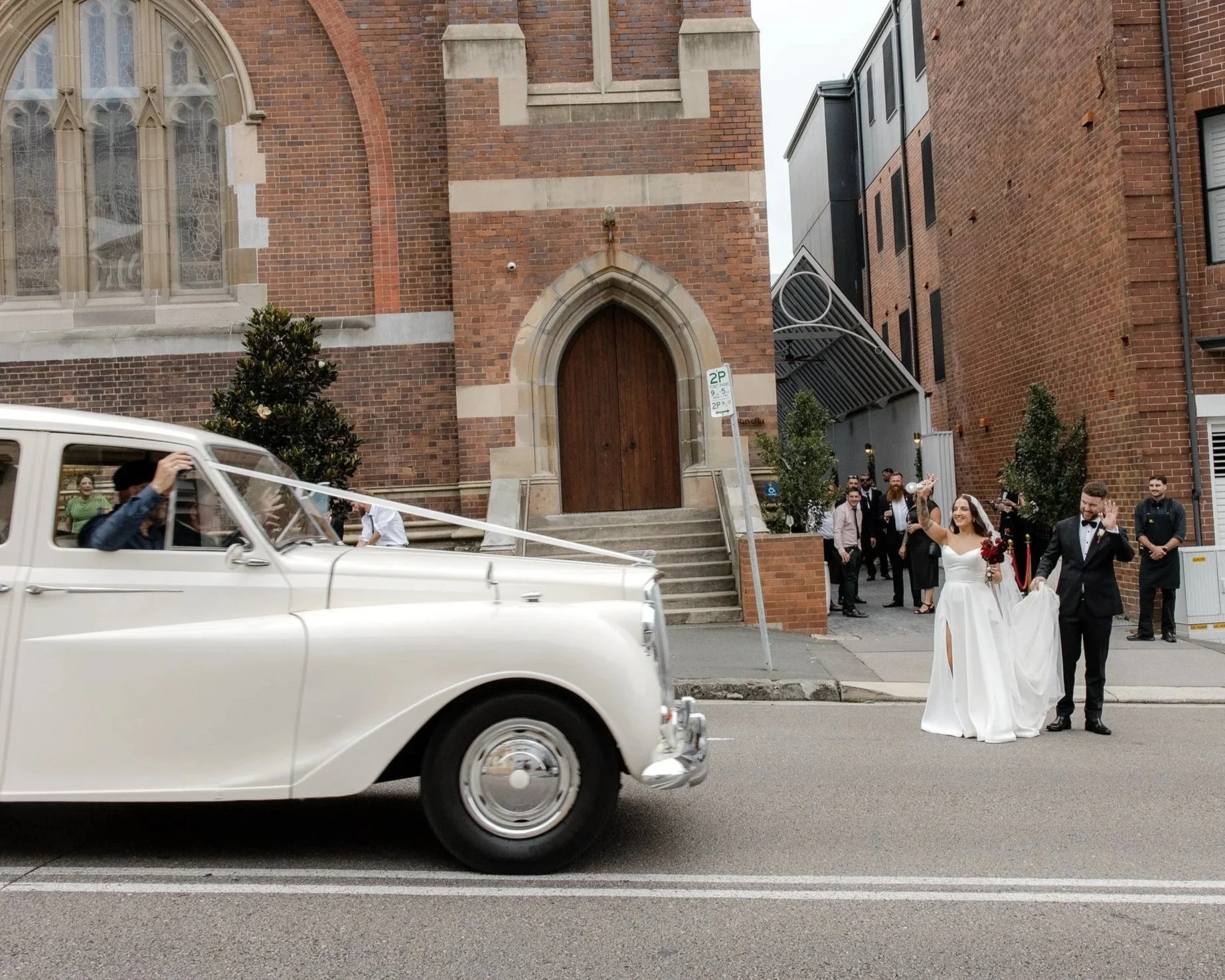 A vintage white car drives past a wedding couple and guests outside a brick church. The bride wears a white gown and veil, holding a bouquet, and the groom is in a black tuxedo. The background features church stairs, brick building, and wedding guests smiling and waving.
