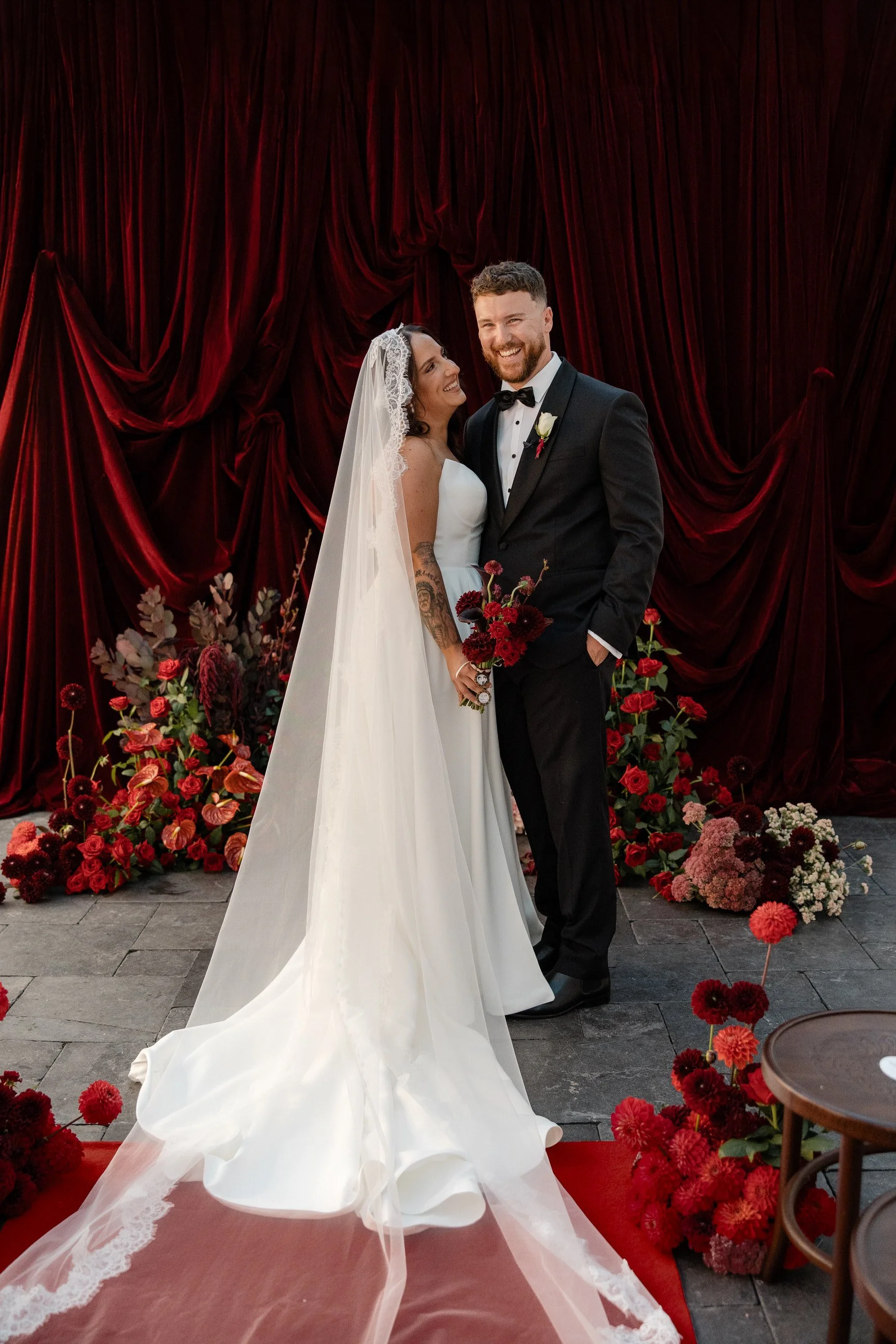 A bride and groom standing in front of a red velvet curtain with floral arrangements, on a red carpeted pathway, during a wedding celebration.