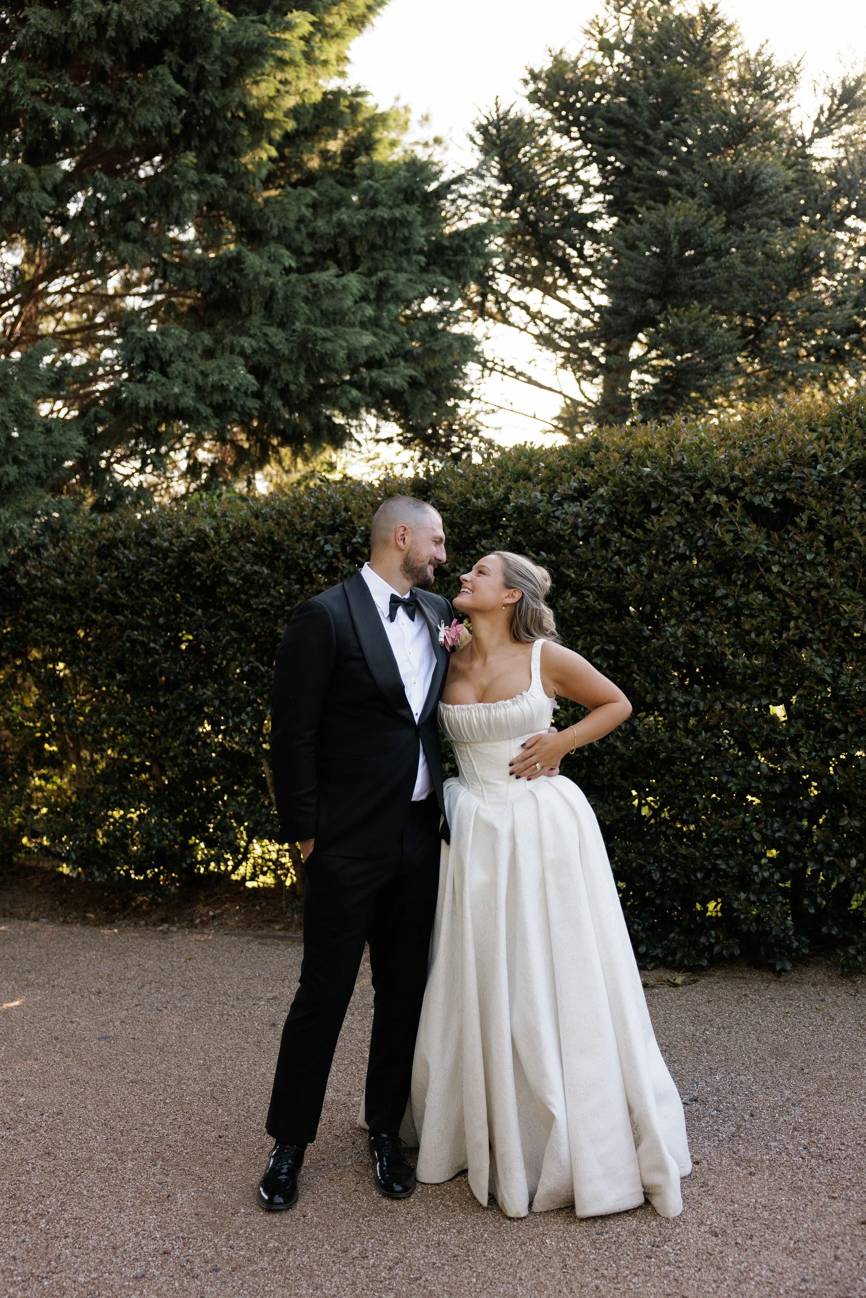 A couple dressed in formal wedding attire, standing outdoors in front of a dense hedge and tall trees with the sun setting behind them. The man is wearing a black tuxedo with a bow tie, and the woman is wearing a white strapless wedding gown. They are smiling and looking at each other.