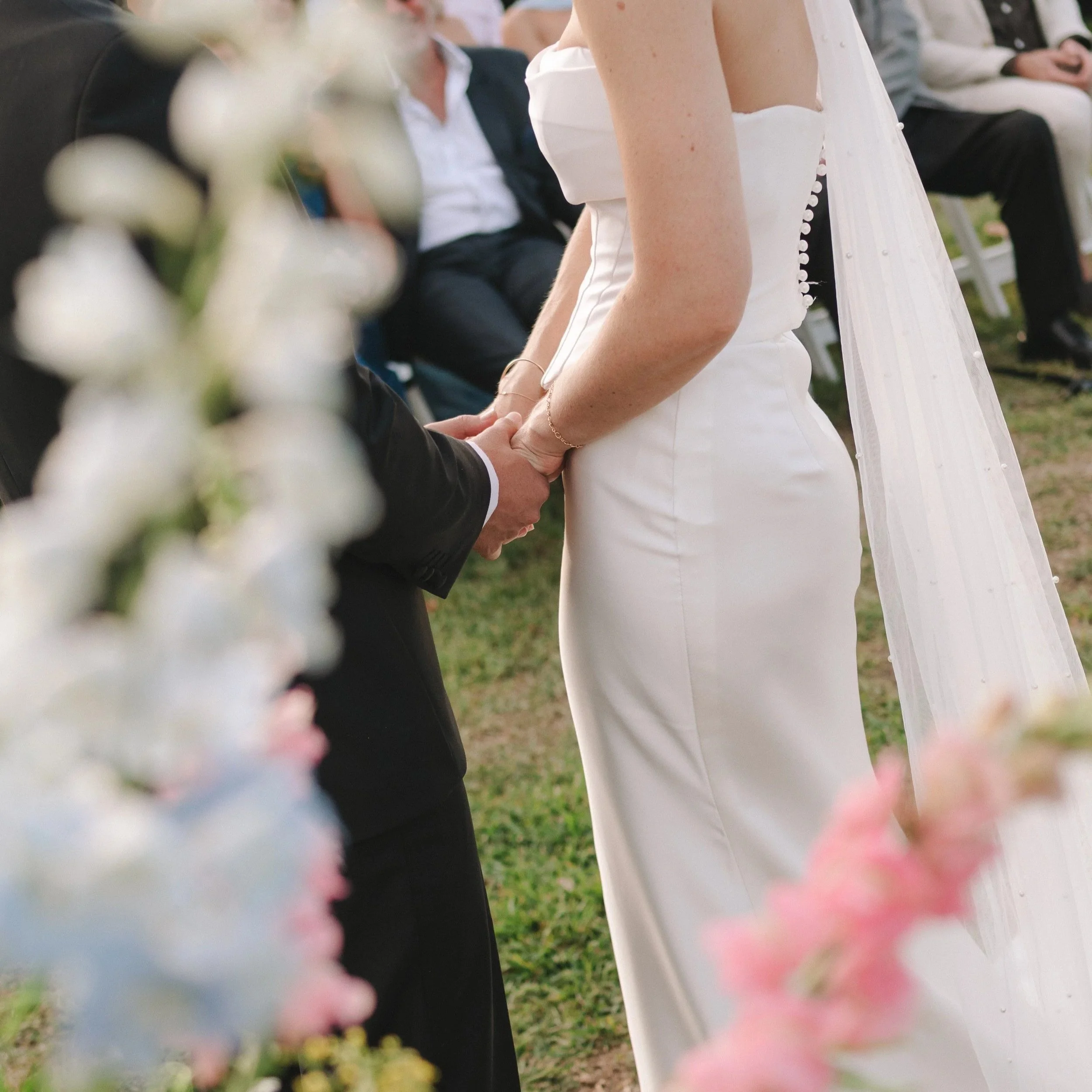 A close-up of a wedding ceremony where a bride and groom are holding hands, with blurred flowers in the foreground and guests in the background.