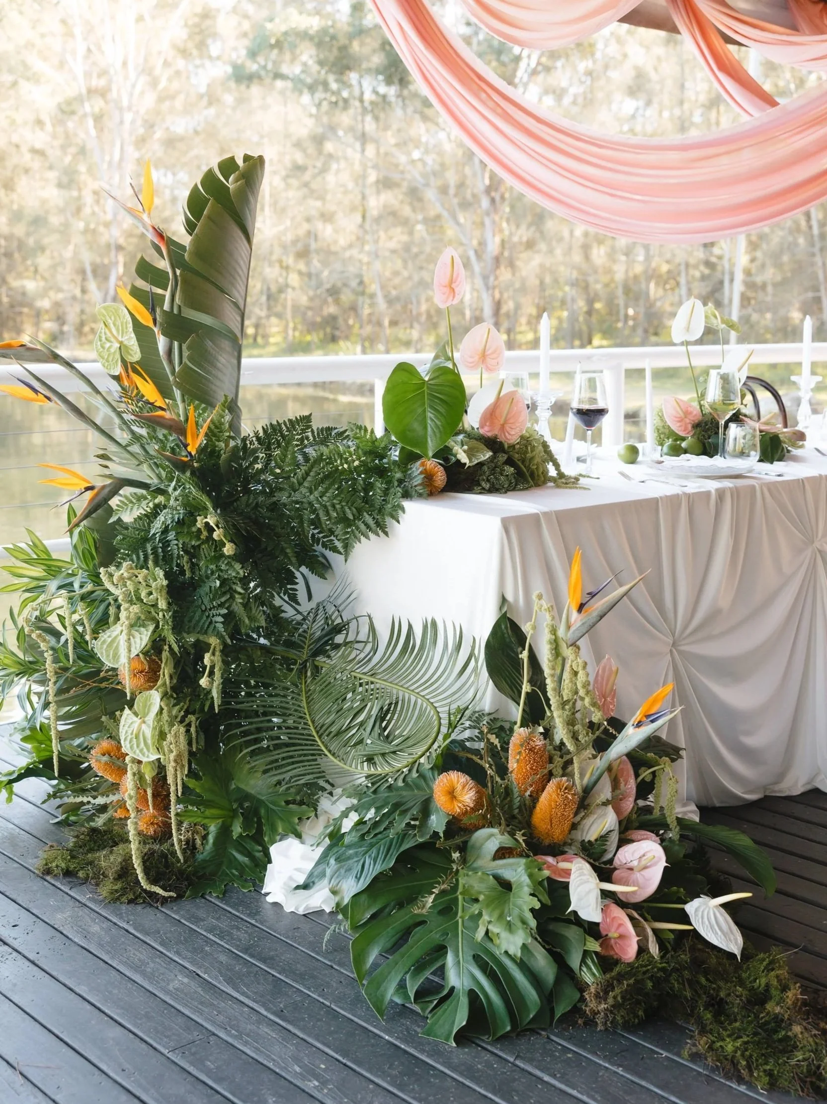 Floral arrangement with tropical plants and flowers on a white tablecloth, surrounded by candles, wine glasses, and greenery, set against a backdrop of trees and draped fabric.