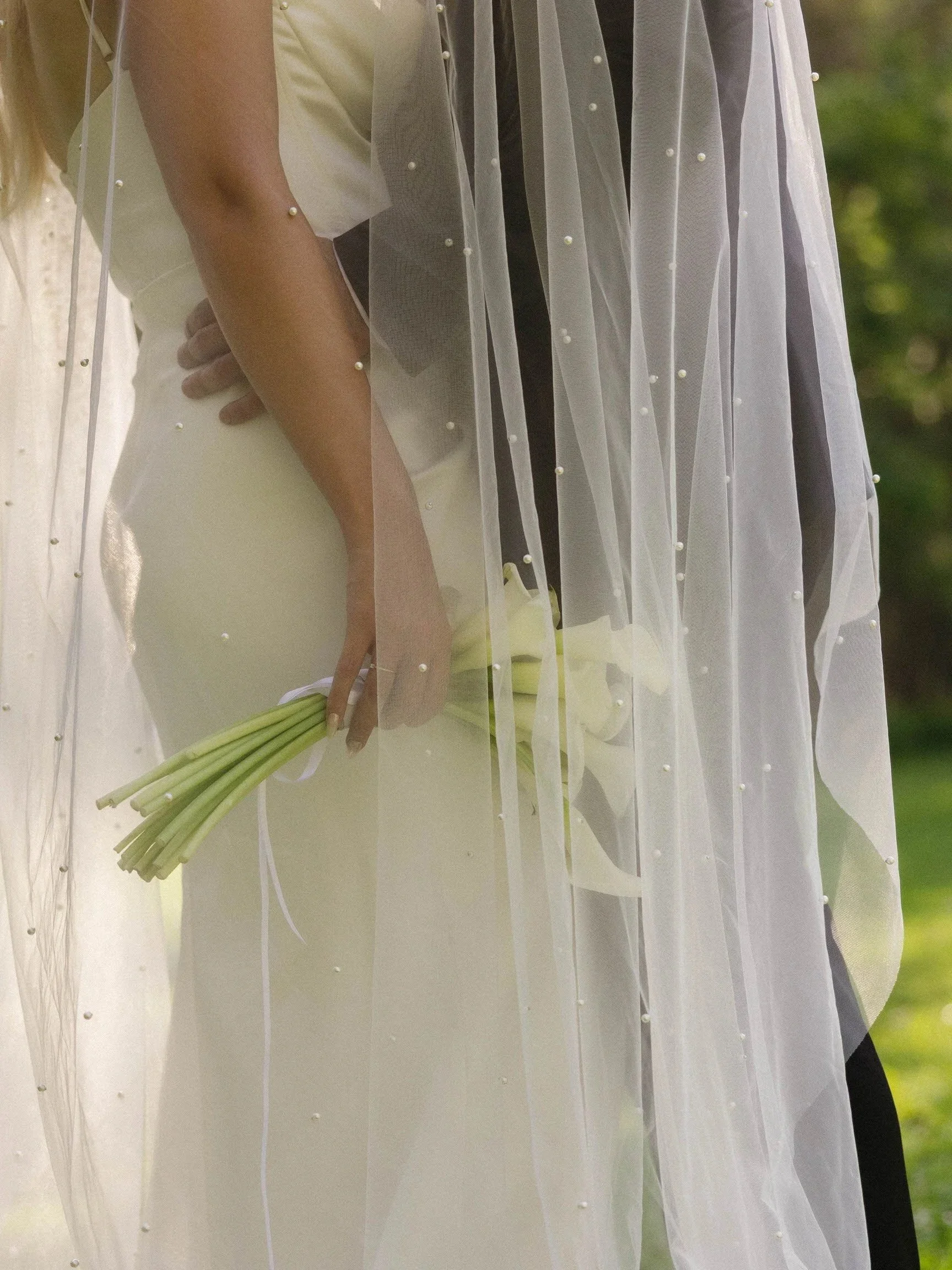 Close-up of a bride holding a bouquet of white calla lilies, wearing a white wedding gown with a sheer veil decorated with small pearls.