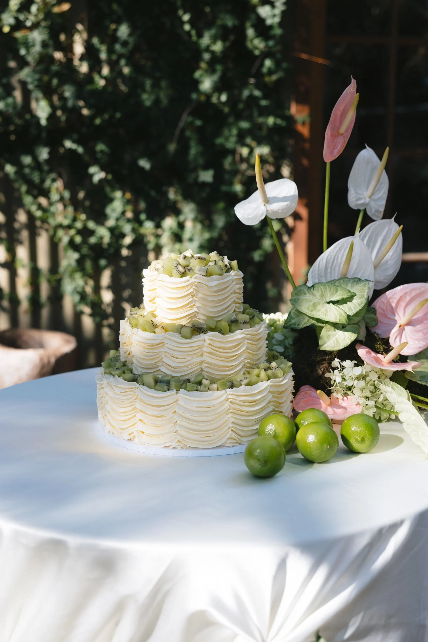 A white tiered cake topped with kiwi slices, decorated with white and pink anthurium flowers, and surrounded by green limes, set on a round white tablecloth outside with greenery in the background.
