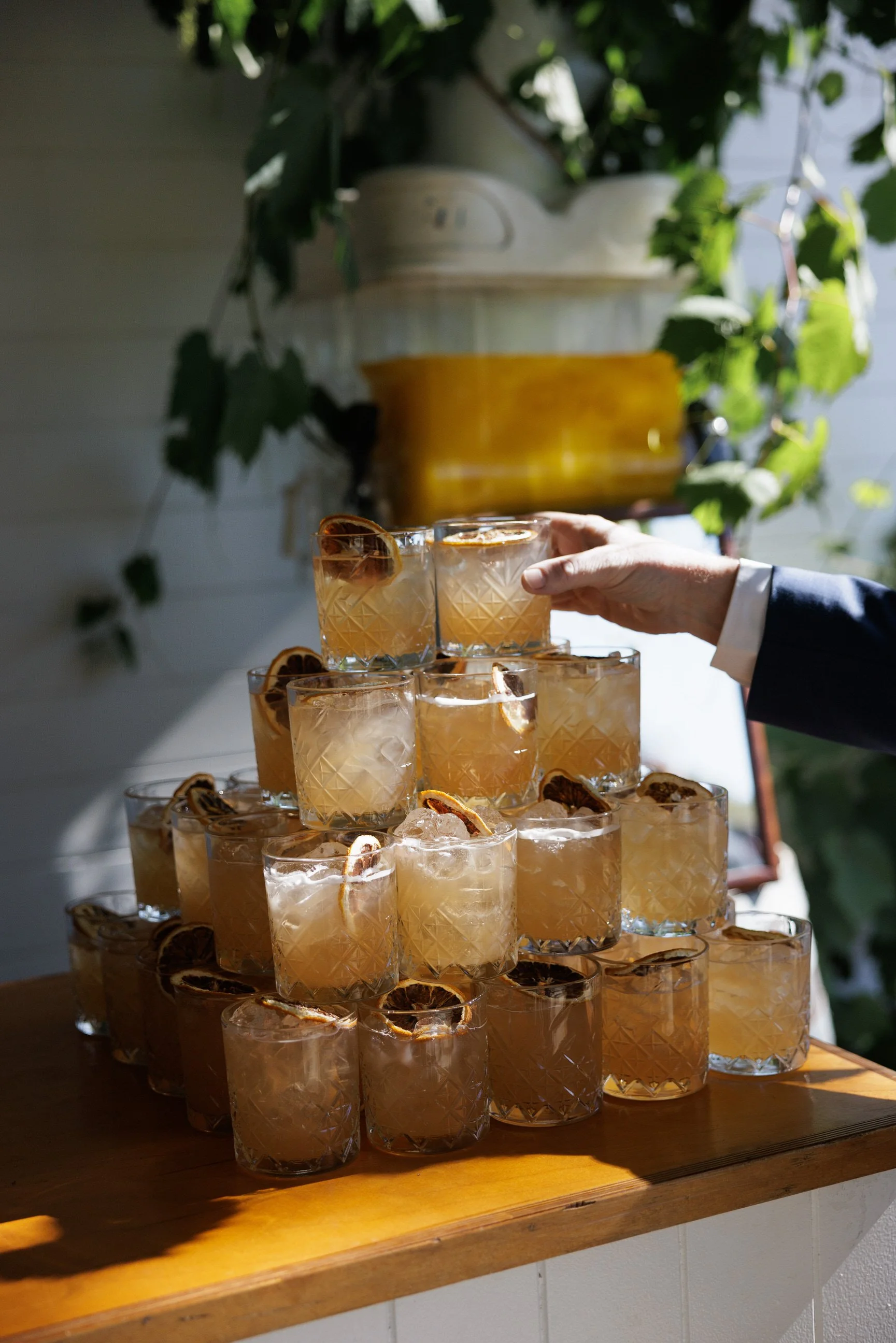 Stacked glasses with iced tea and lemon slices on a wooden surface, with greenery and a white house in the background.