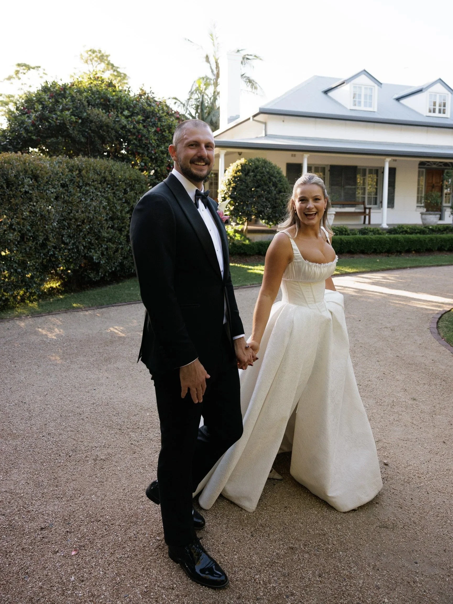 A happy couple in wedding attire standing outside in front of a large house and garden, holding hands and smiling at the camera.