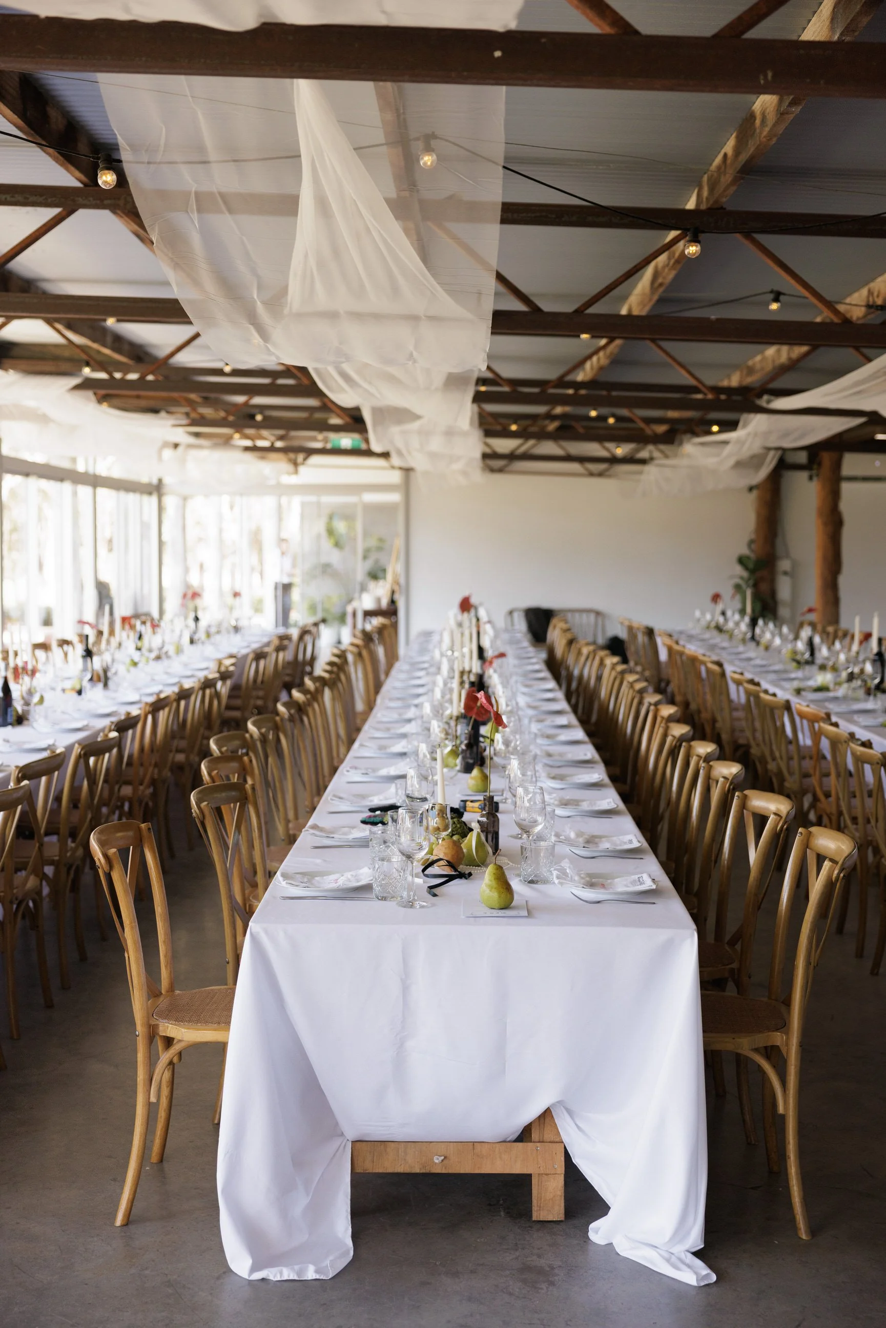 Long banquet table with white tablecloth, set with wine glasses, plates, silverware, and small flower arrangements. Wooden chairs surround the table. Overhead, sheer white fabric drapes hang from the exposed wooden beams of a high-ceiling venue. In the background, large windows let in natural light, and there are some plants against the white walls.