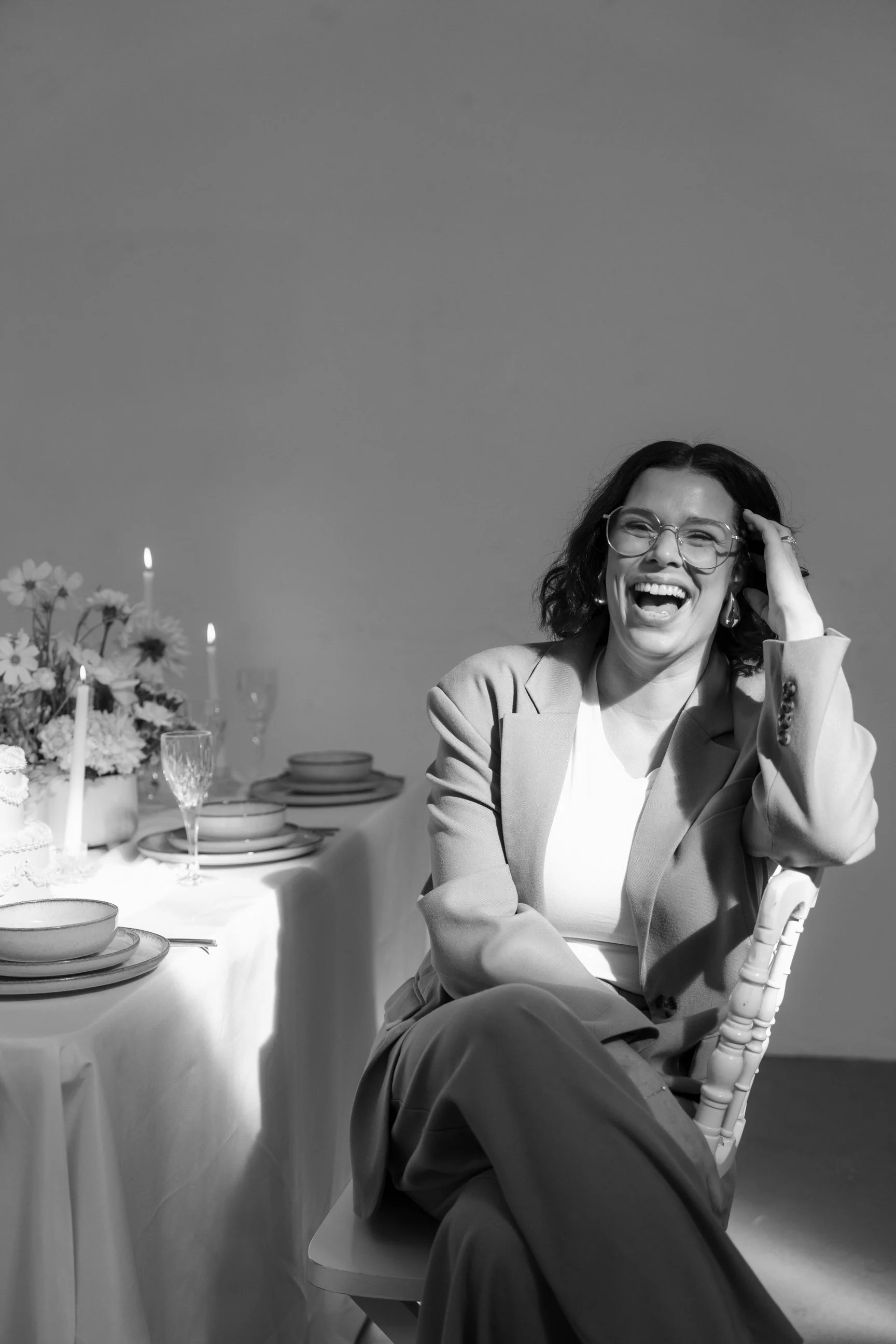 A woman with glasses, short curly hair, and a blazer, sitting at a dinner table, laughing with her hand on her head, in black and white.