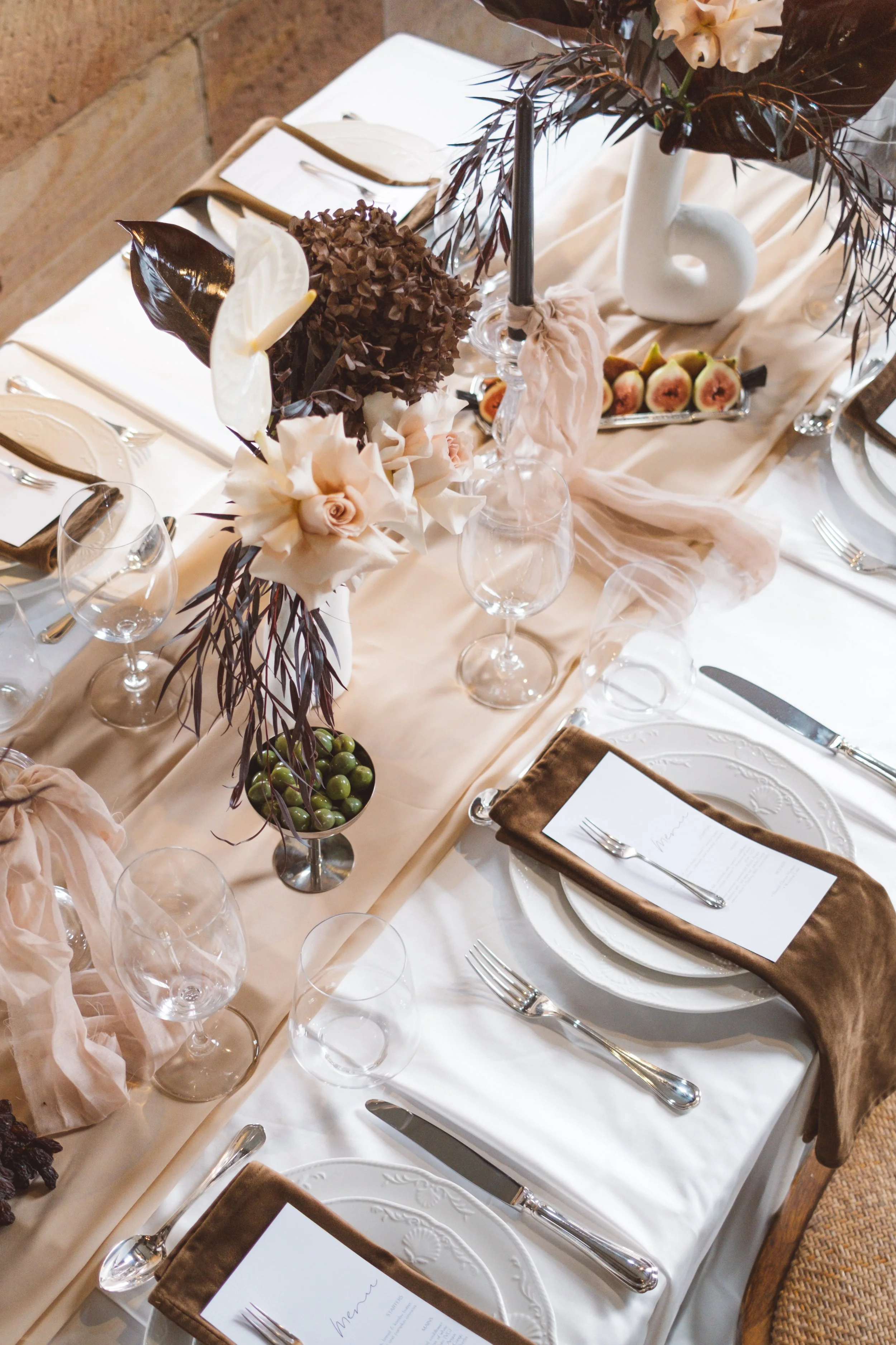 A formal dining table decorated with elegant floral arrangements and set with plates, silverware, wine glasses, and napkins for a wedding reception