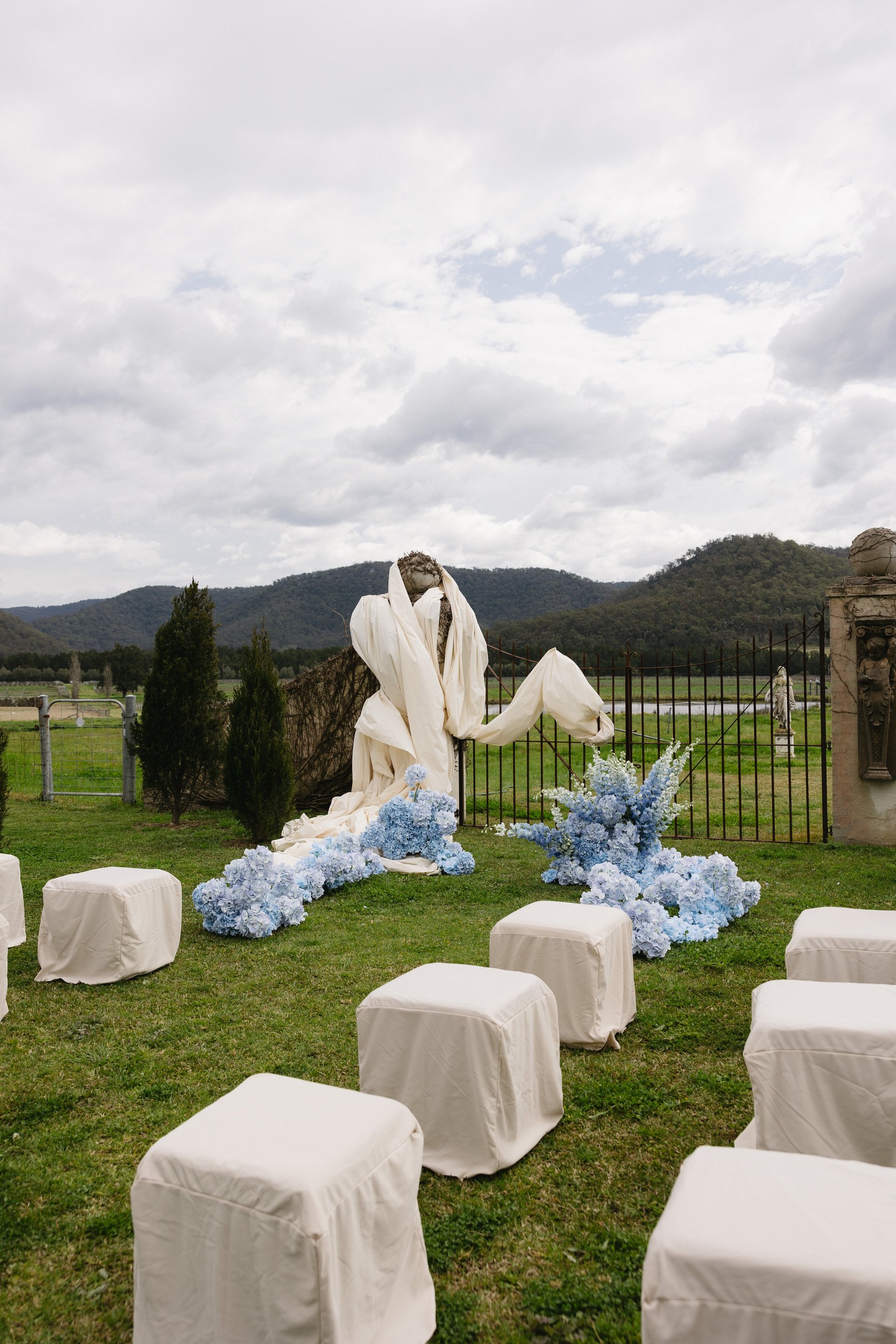 An outdoor wedding setup with white chairs covered in fabric, blue and white floral arrangements, and a draped fabric backdrop against a rural landscape with mountains and a cloudy sky.