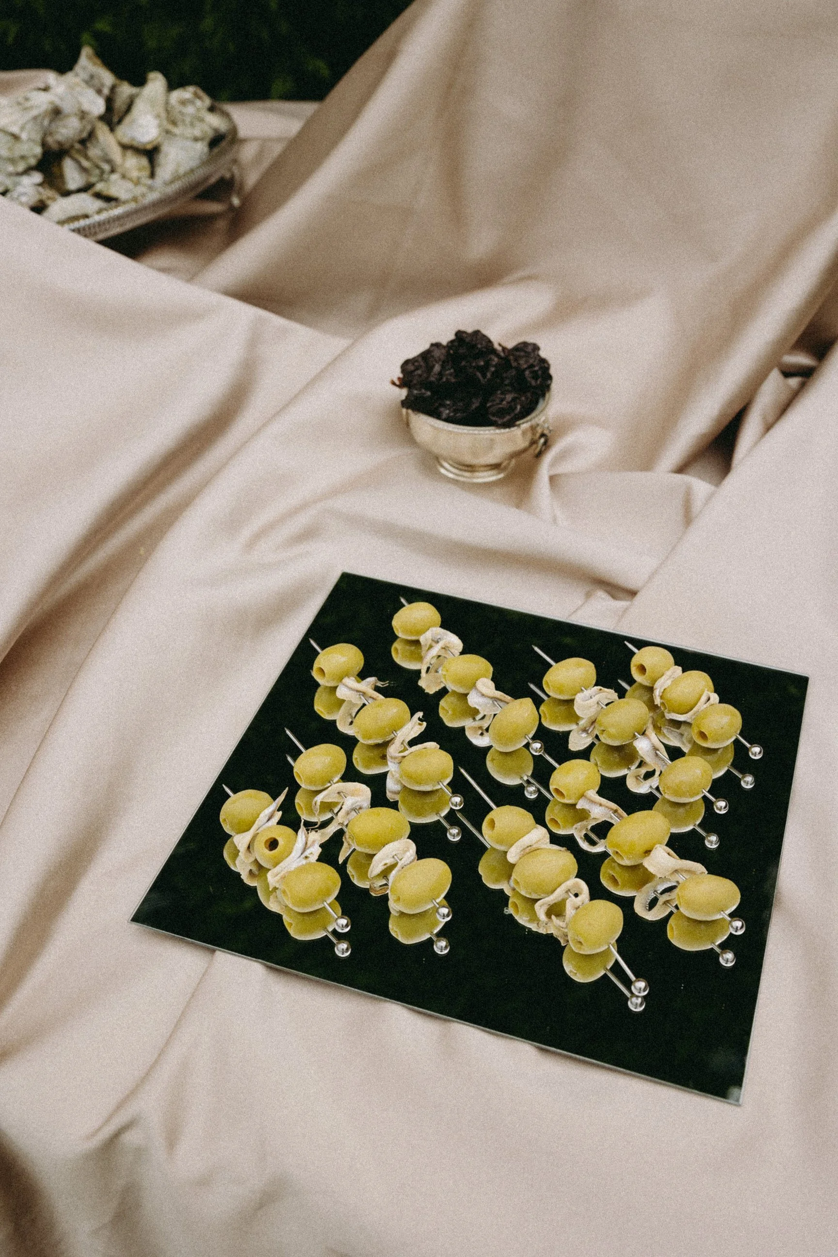 A close-up of a display of skewers with yellow grapes and shells along a black surface, with a silver bowl of dark dried fruit and a tray of Doberge cookies in the background, all placed on a satin fabric surface.