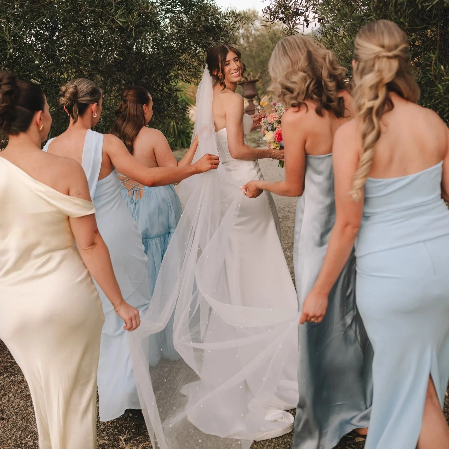 A smiling bride in a white wedding dress holding a bouquet of colorful flowers, surrounded by bridesmaids in pastel dresses, outdoors among trees.