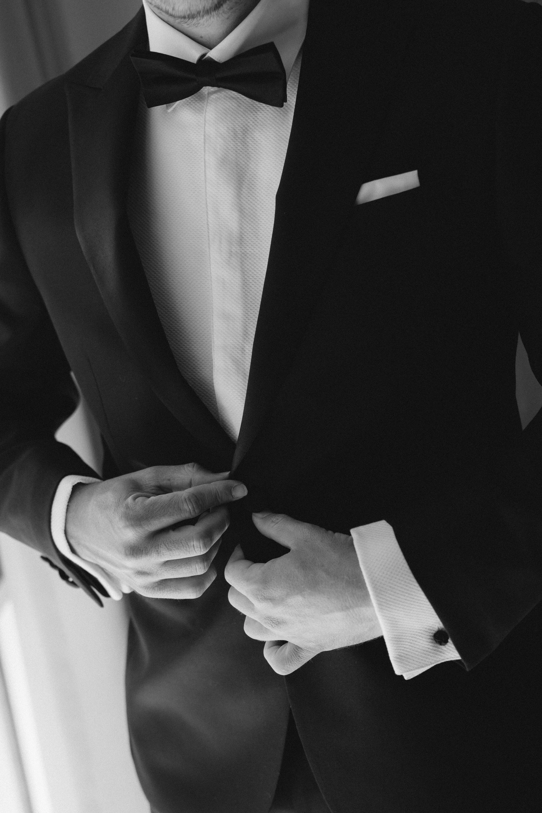 Close-up of a man adjusting his tuxedo jacket, wearing a white shirt and black bow tie, with a white pocket square visible.