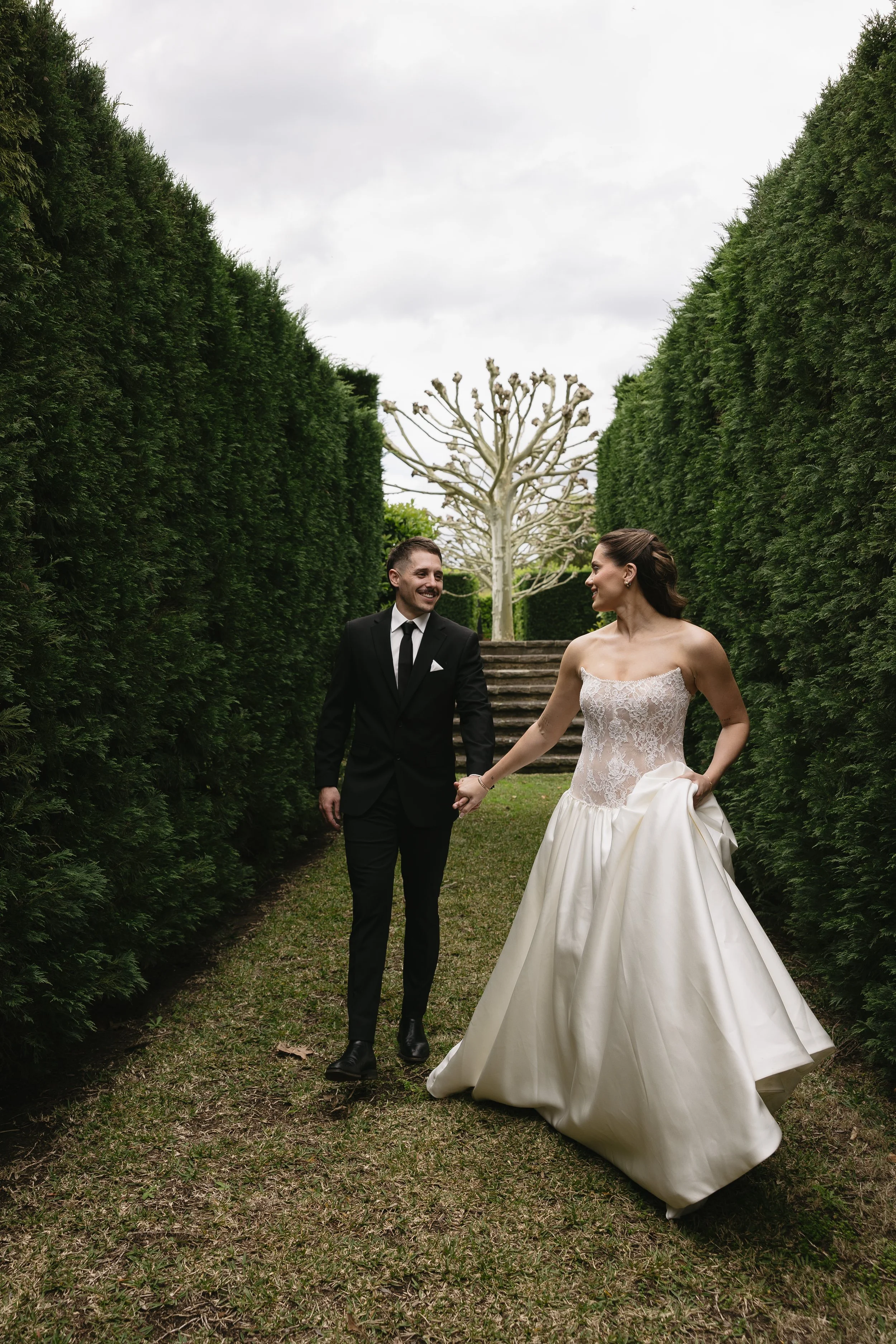 A bride and groom walking hand in hand in a garden pathway surrounded by tall green hedges, with a leafless tree in the background, under an overcast sky.