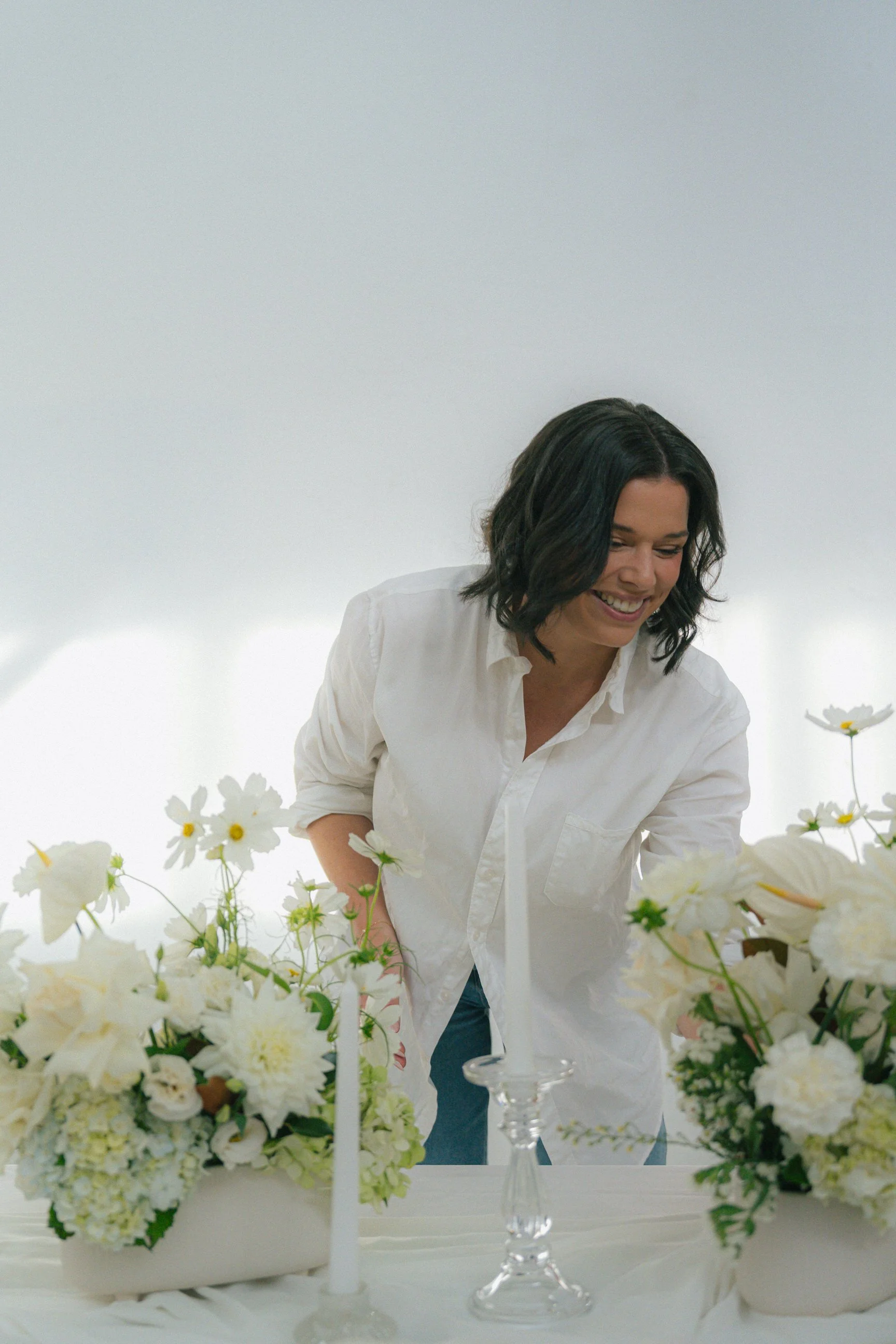 Woman with short dark hair wearing a white shirt, smiling, surrounded by white flowers and candles on a white table in a bright space.