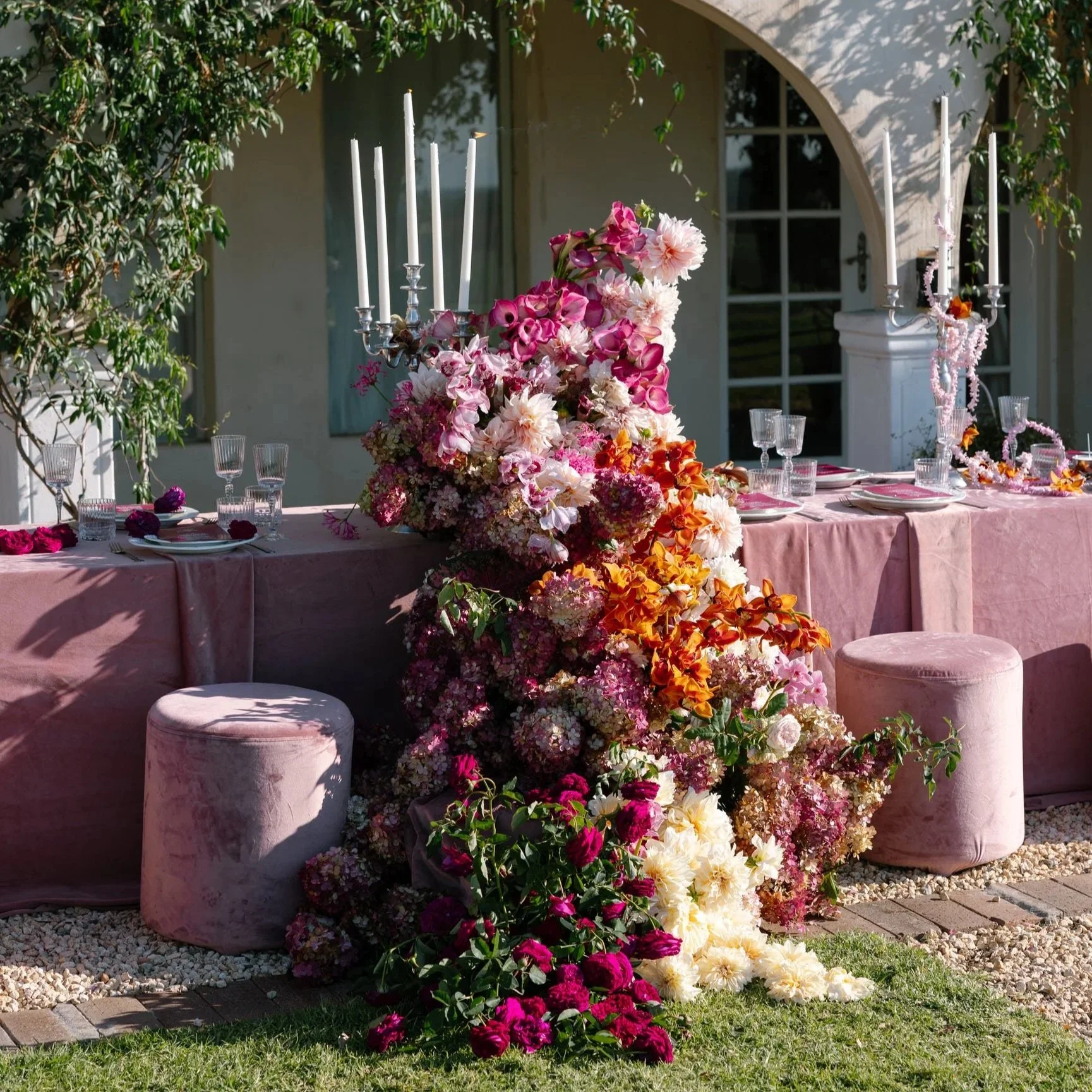 Elegant outdoor table setting with a long pink tablecloth, large central floral arrangement with pink, white, and orange flowers, surrounded by pink velvet stools, and decorated with white candlesticks and crystal glasses. A lush green tree and a house with white walls and black-framed windows are in the background.